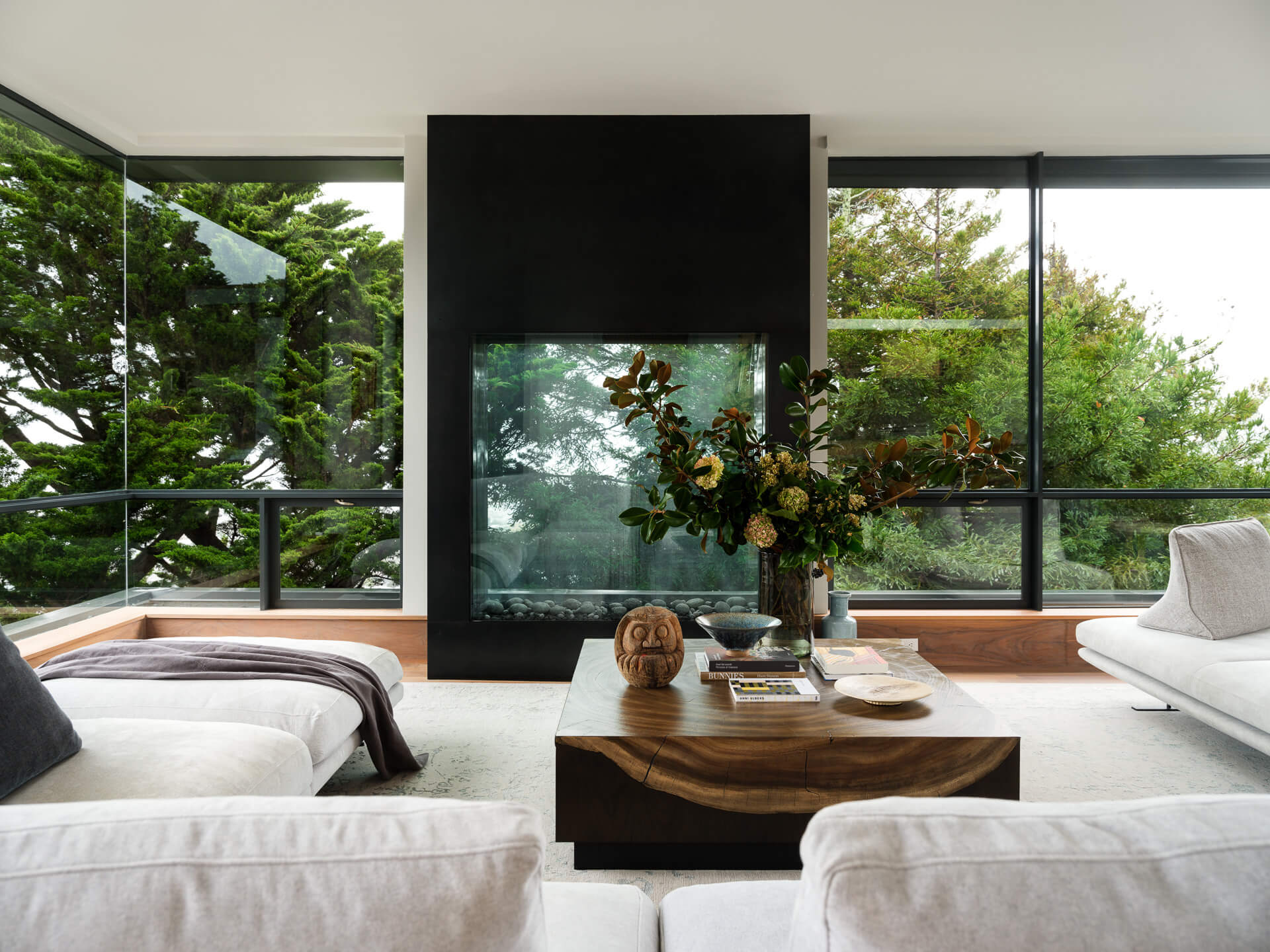 Contemporary living room with white seating, wood coffee table, central black fireplace, and leafy views.