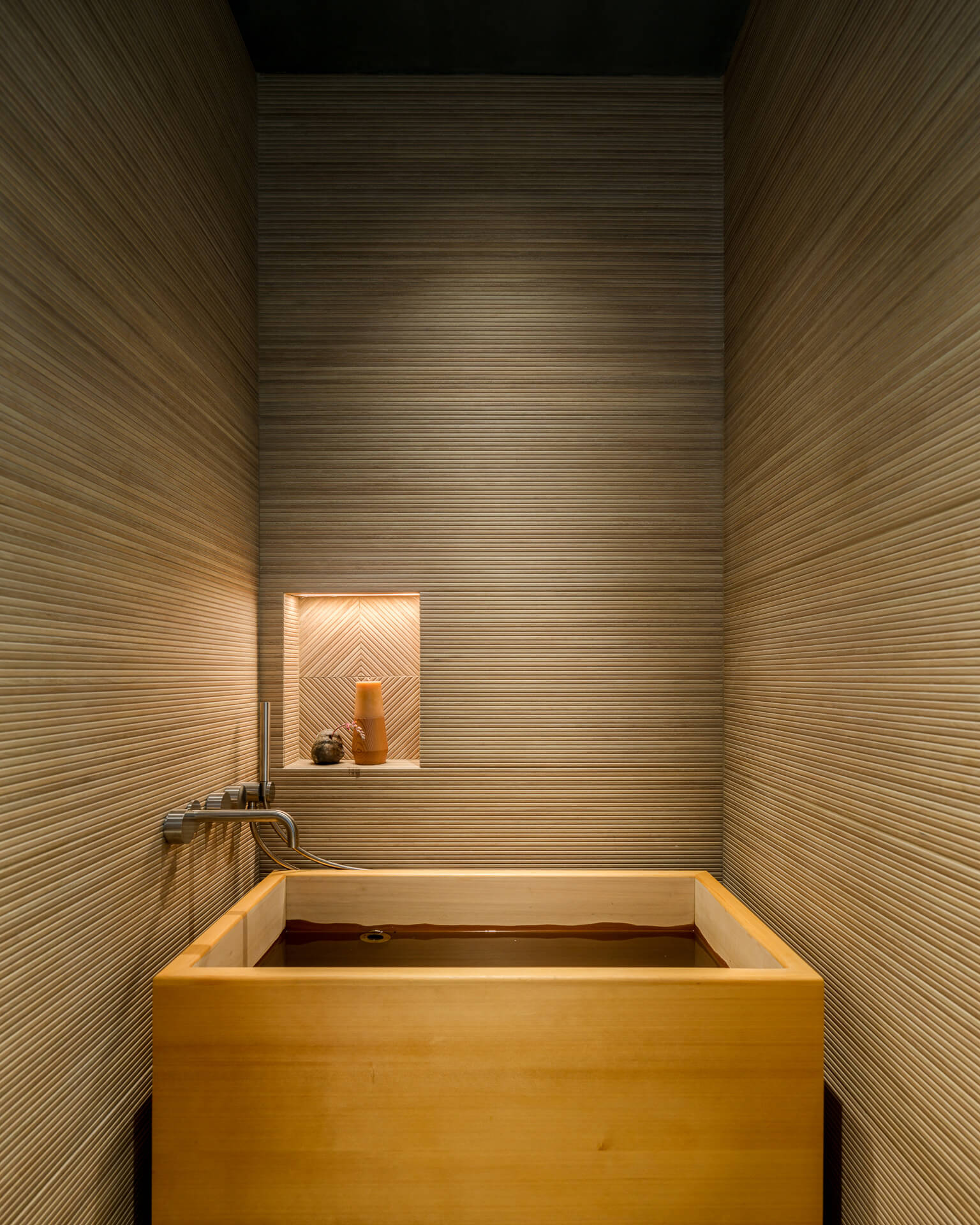 Japanese-style wood soaking tub in a narrow wood-paneled room with a softly lit recessed niche.