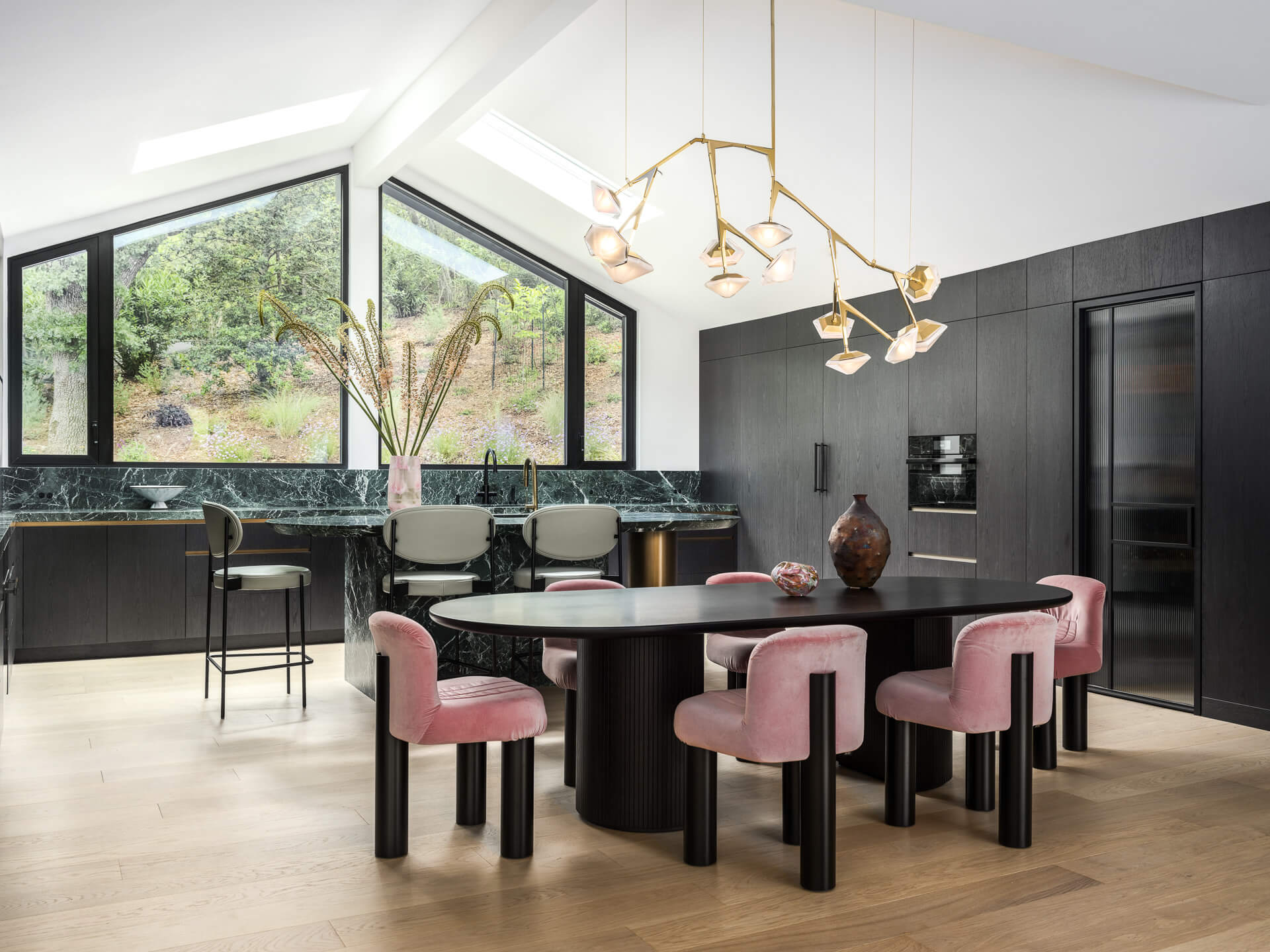Contemporary kitchen and dining area with black cabinetry, pink dining chairs, sculptural chandelier, and large windows overlooking greenery.