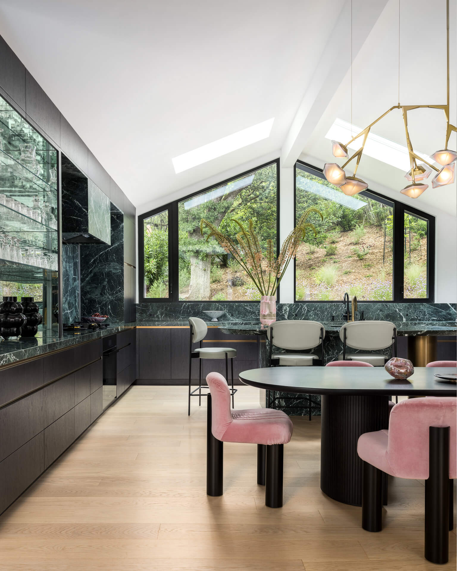 Contemporary kitchen and dining area with black cabinetry, pink dining chairs, sculptural chandelier, and large windows overlooking greenery.