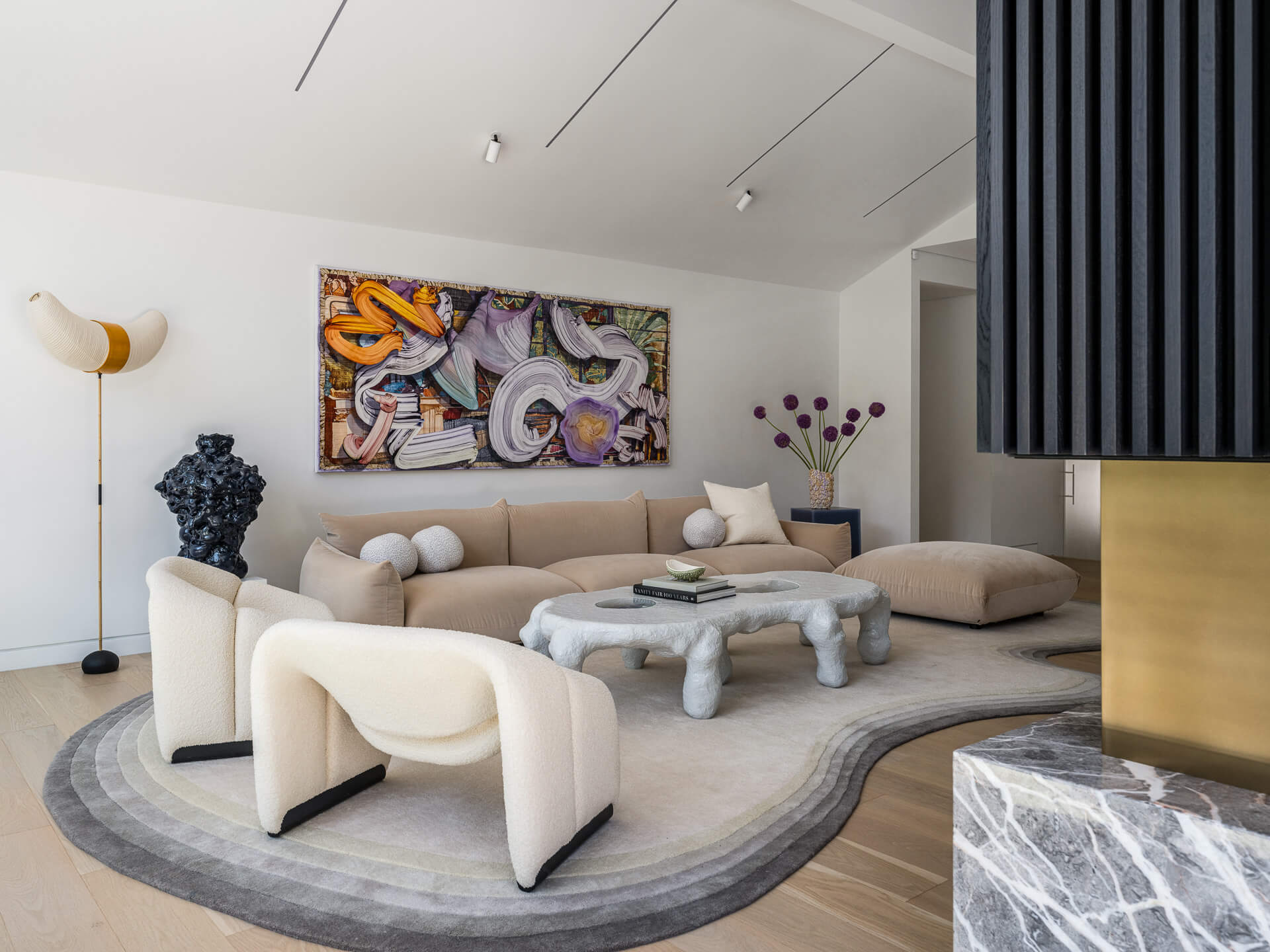 Bright living room with curved cream seating, sculptural coffee table, colorful artwork, and layered circular rug.