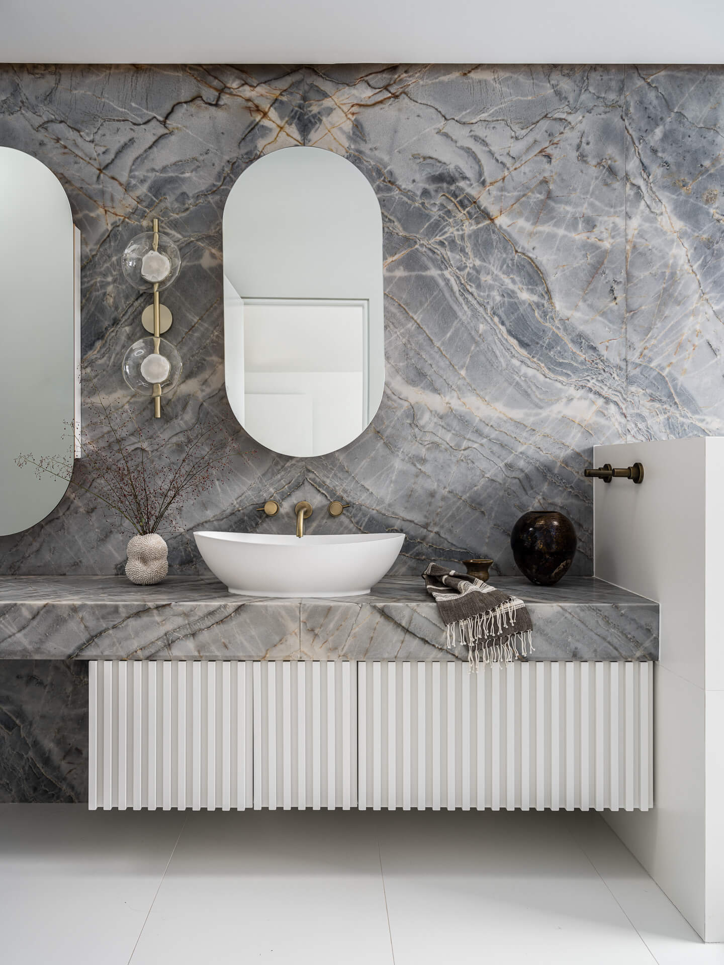 Marble bathroom vanity with vessel sink, rounded mirror, white ribbed cabinetry, and wall-mounted lighting.
