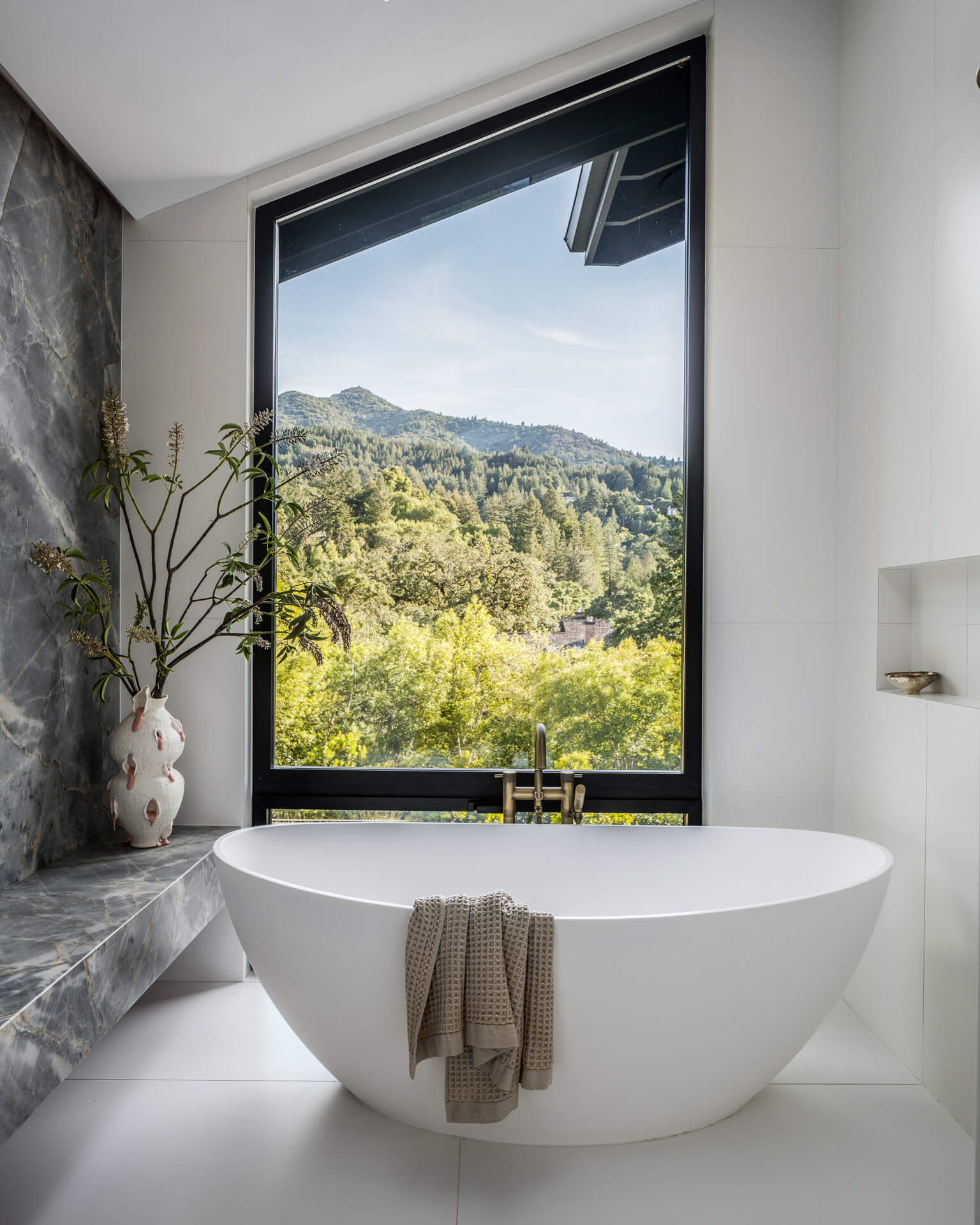 Freestanding white bathtub beside a large picture window overlooking wooded hills.