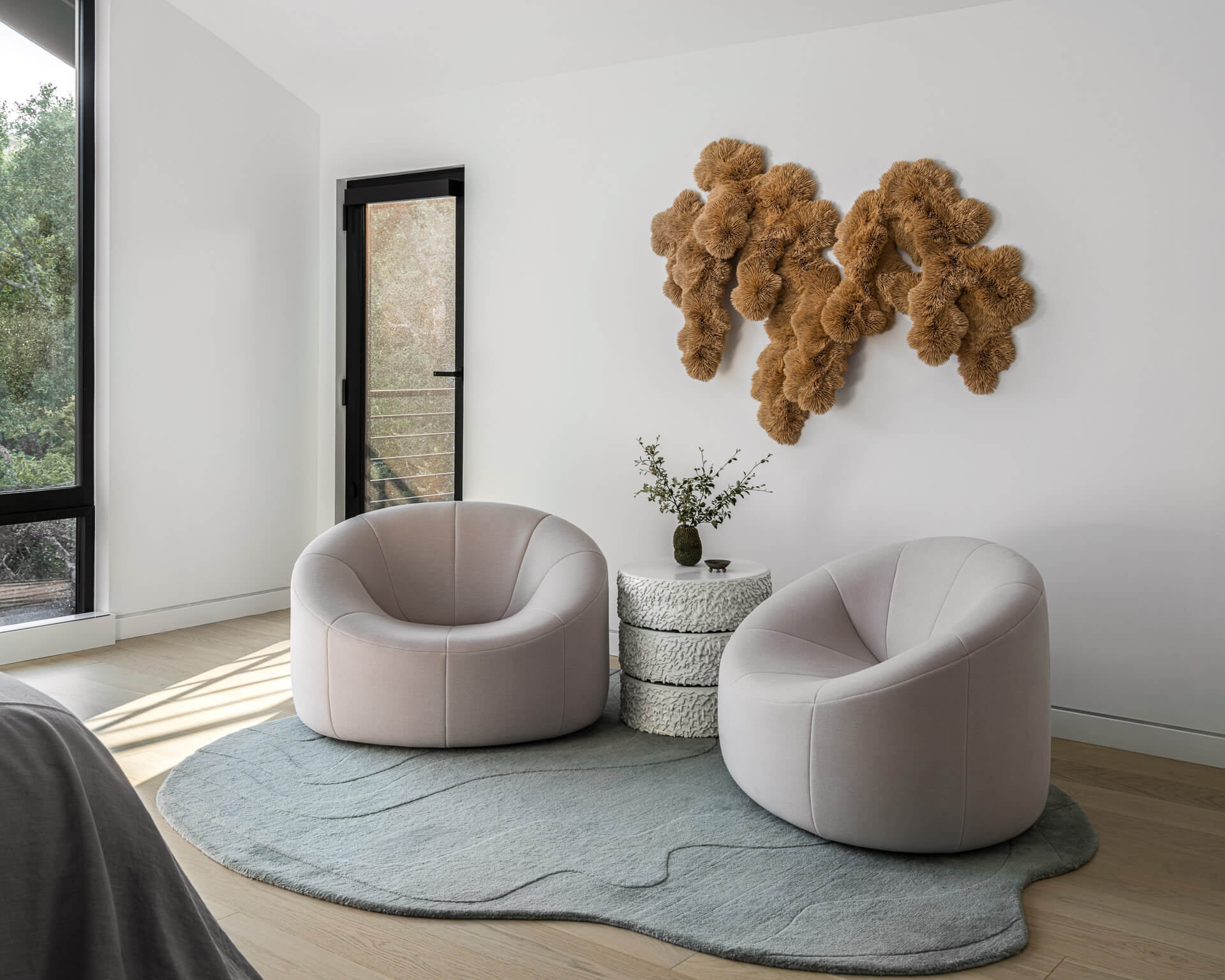 Sitting area with two rounded lounge chairs, pale sculptural rug, narrow window, and textured wall sculpture.