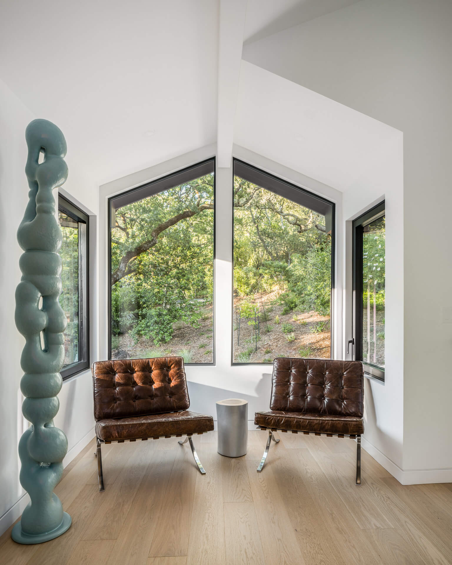 Two brown leather lounge chairs beside a tall blue sculpture and corner windows overlooking trees.