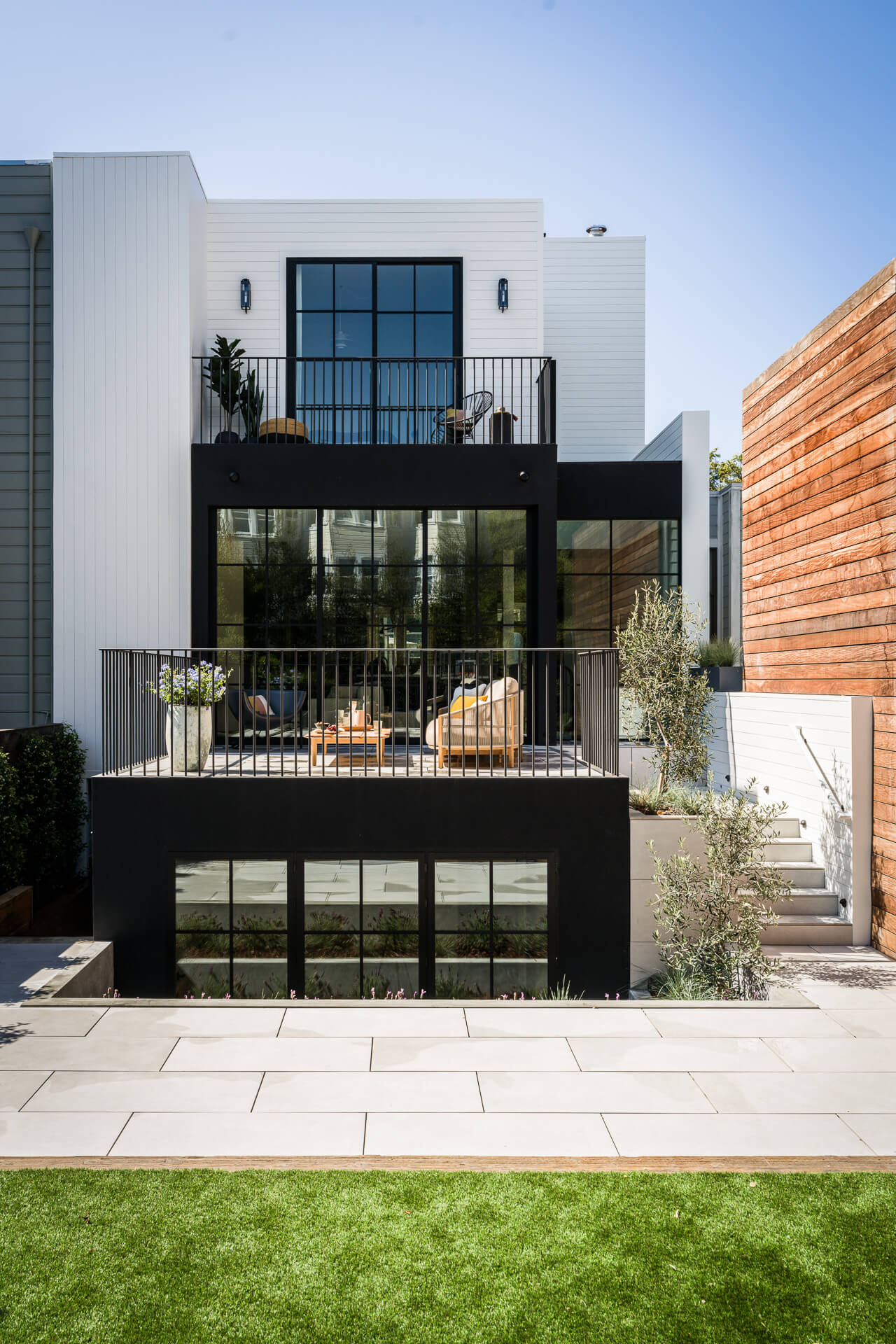 Rear view of a white home with black-framed windows, stacked terraces, stone patio, and lawn.