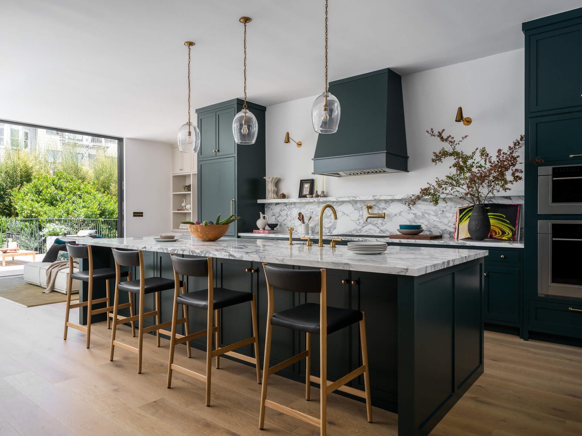 Kitchen with deep teal cabinetry, marble island, black-and-brass stools, and glass pendant lights.