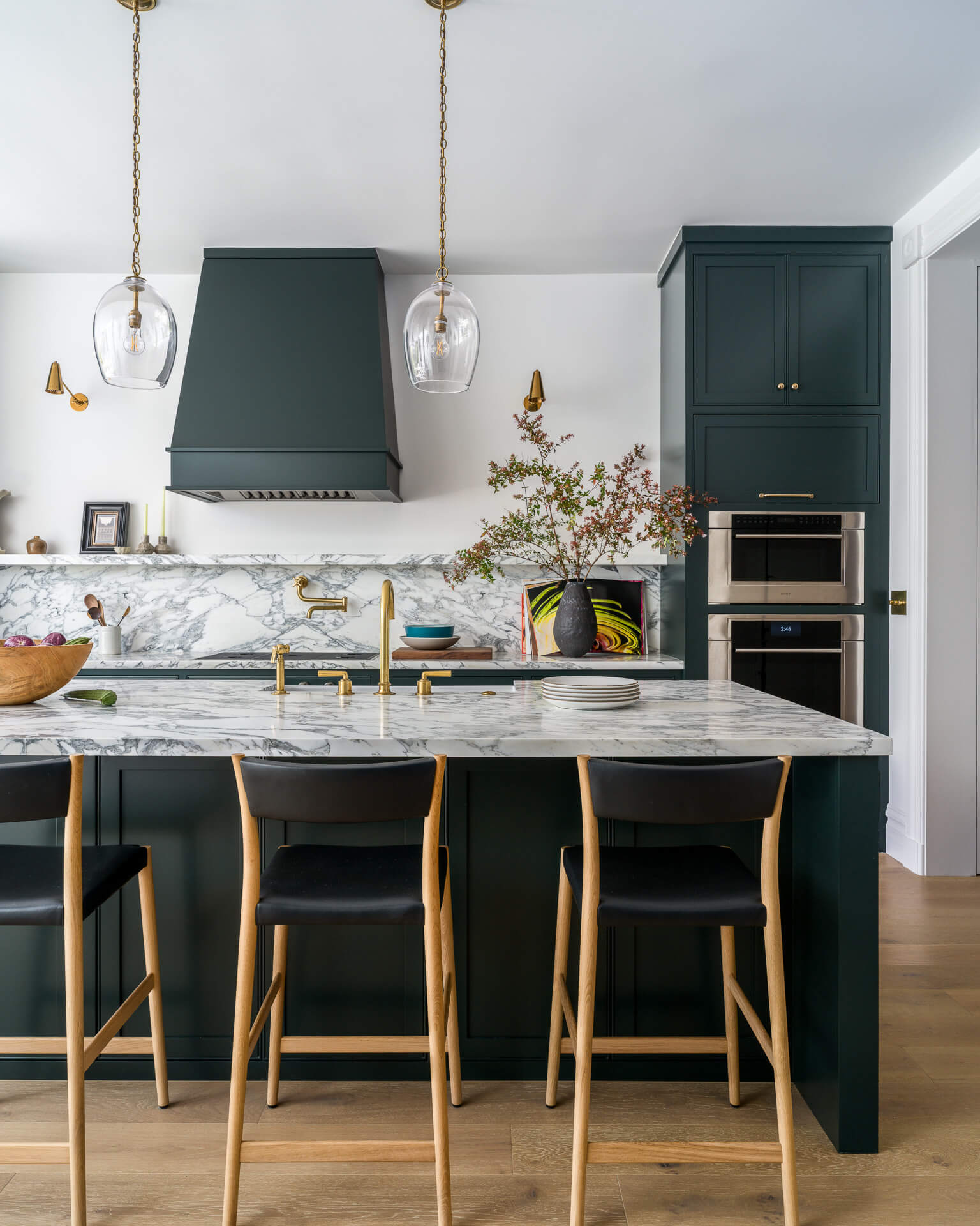 Deep teal kitchen with marble island and backsplash, brass fixtures, and three counter stools.
