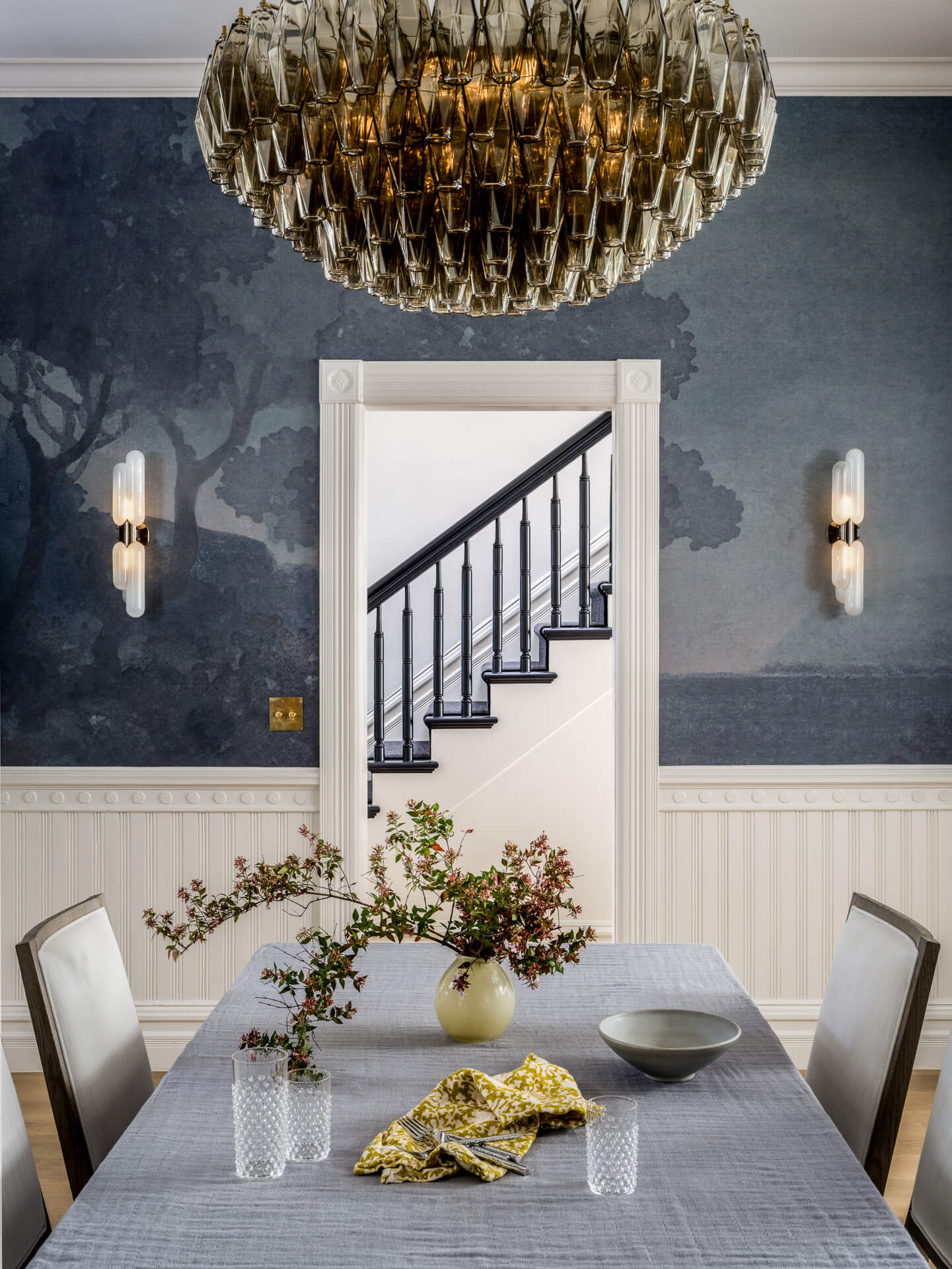 Dining room with blue scenic wallpaper, dramatic glass chandelier, and a gray-covered table.