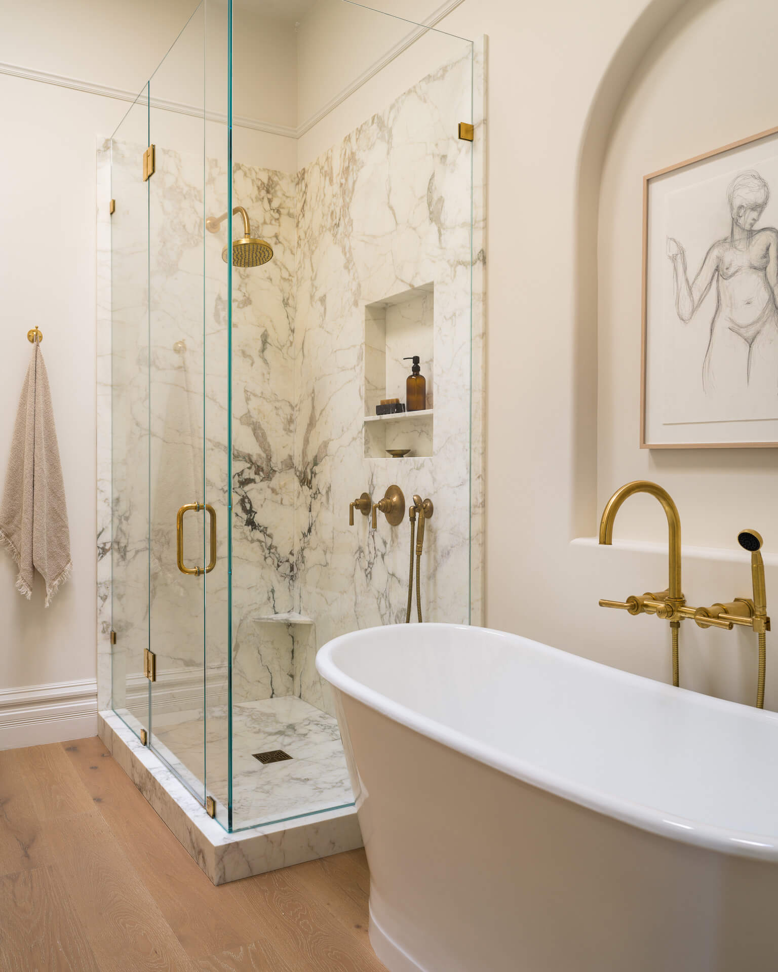 Marble bathroom with freestanding white tub, glass-enclosed shower, brass fixtures, and light wood floors.