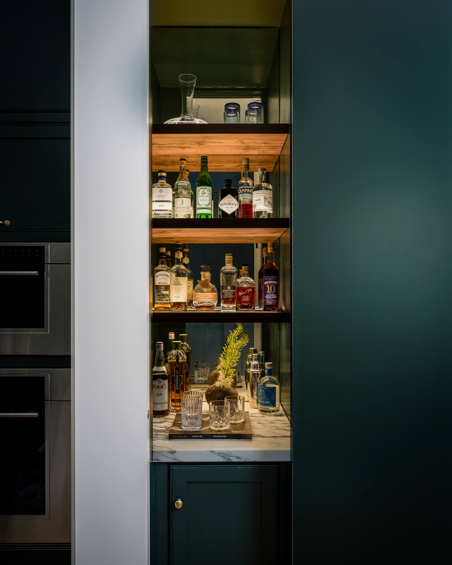 Concealed home bar with deep teal cabinetry, wood shelves, marble counter, and neatly arranged bottles.