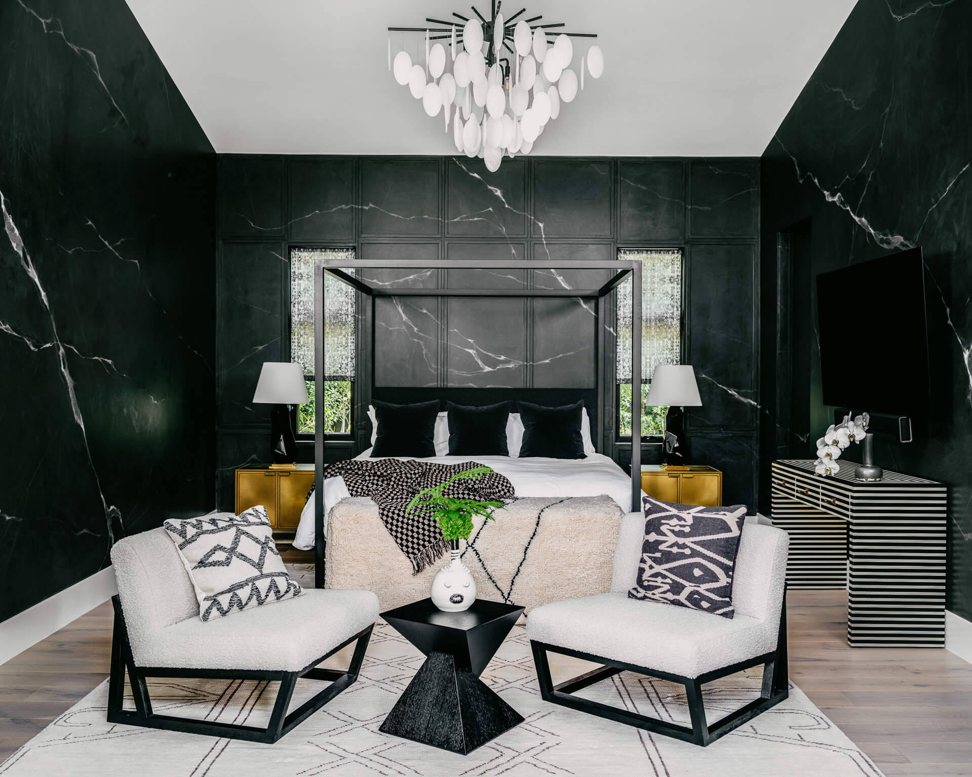 Dramatic bedroom with black marble walls, black canopy bed, patterned lounge chairs, and sculptural chandelier.