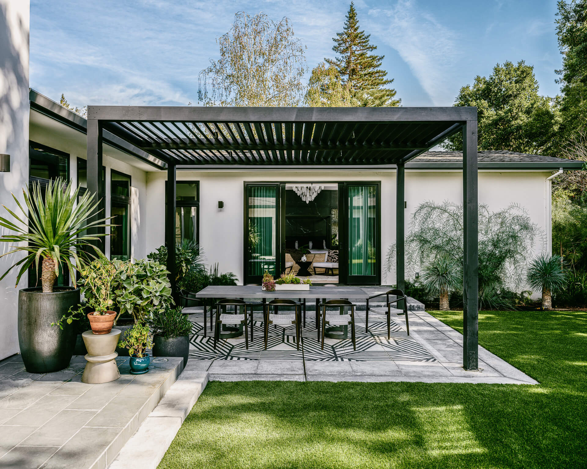 Outdoor dining terrace beneath a black pergola, surrounded by lawn and lush garden plantings.