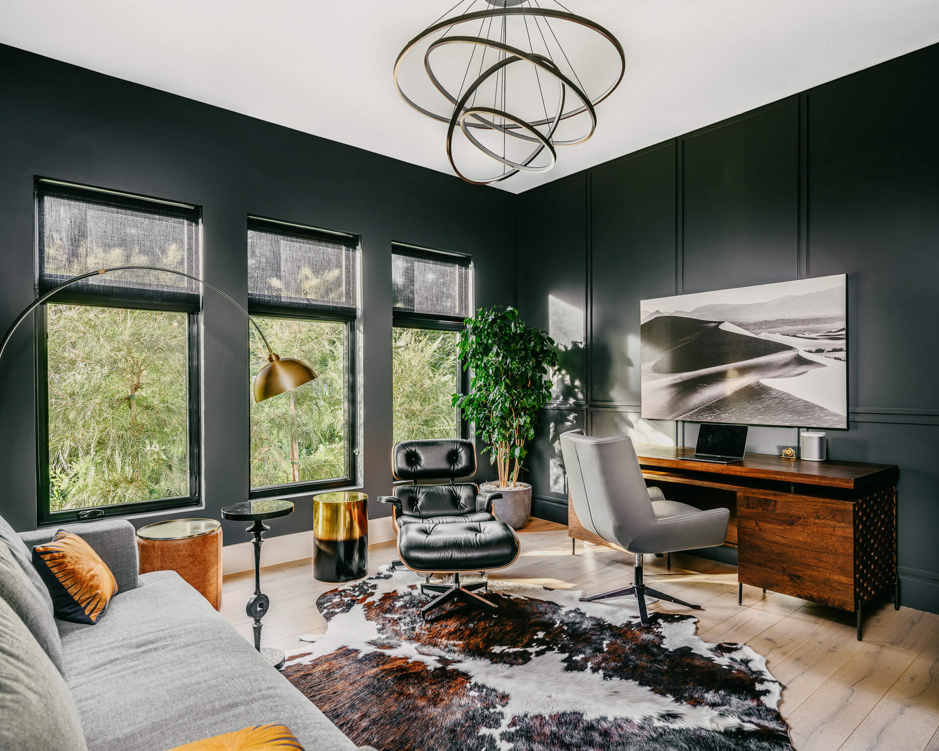 Dark green sitting room with large windows, gray seating, wood desk, and layered artwork.