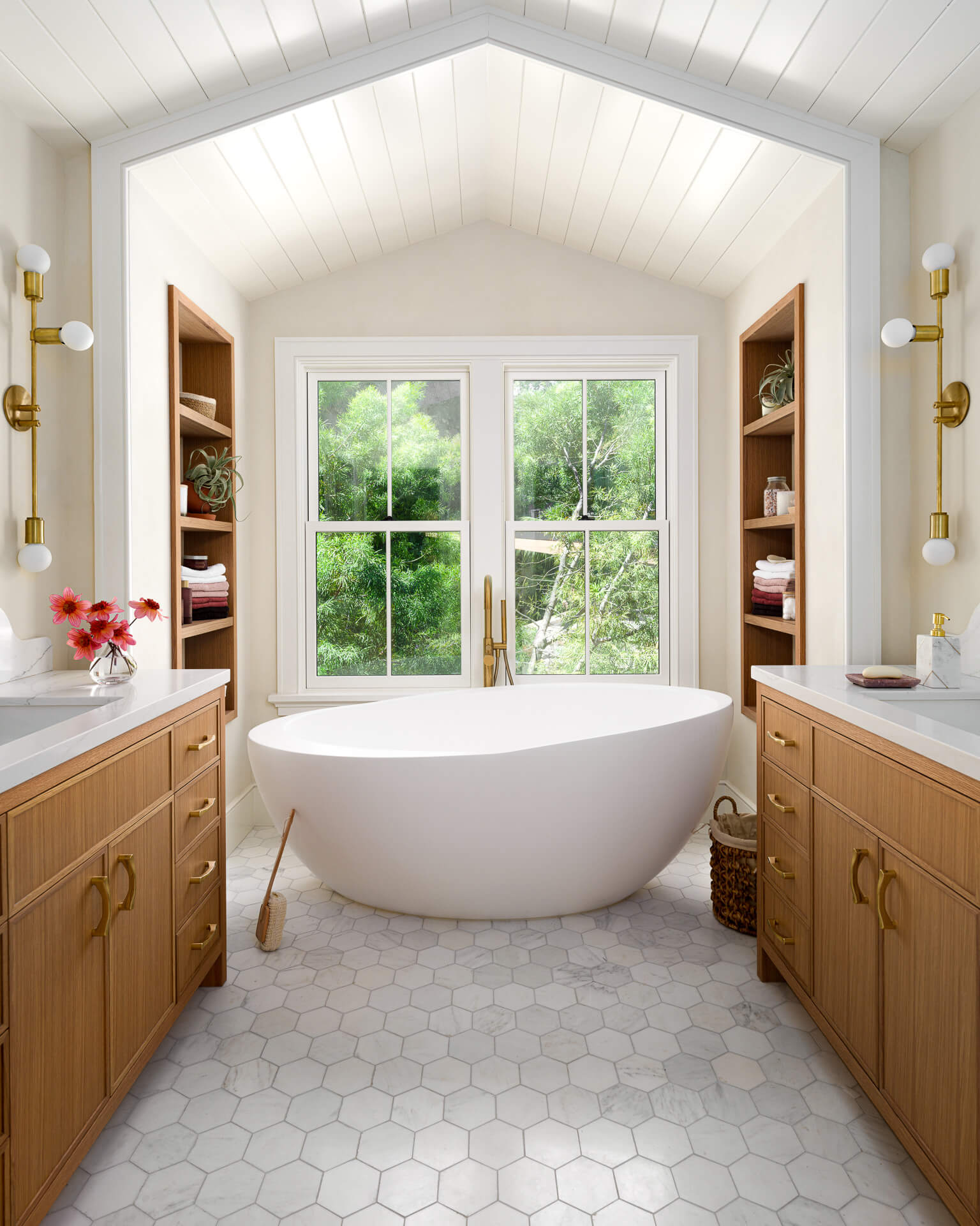 Bright bathroom with freestanding white tub, wood vanities, brass fixtures, and vaulted ceiling.