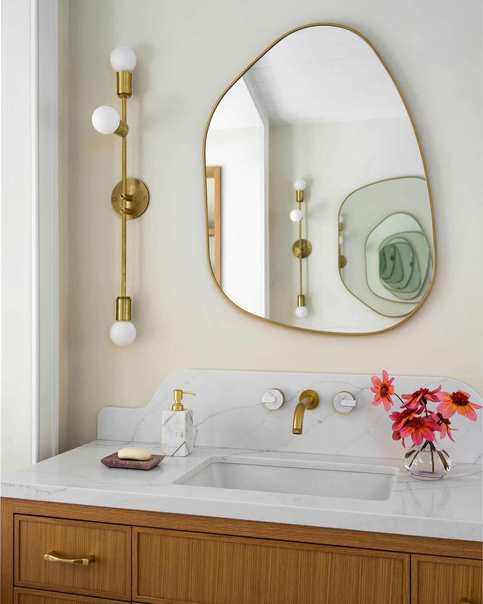 Wood bathroom vanity with white countertop, curved brass mirror, globe sconces, and red flowers.