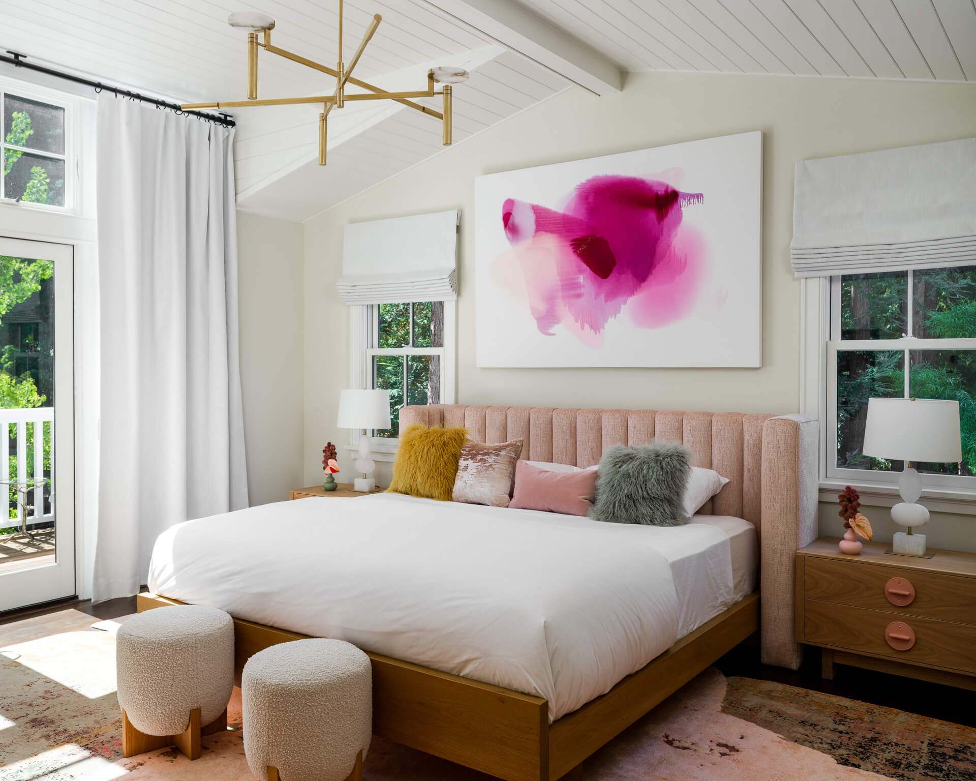 Sunlit bedroom with wood bed, blush headboard, colorful pillows, abstract artwork, and balcony doors.