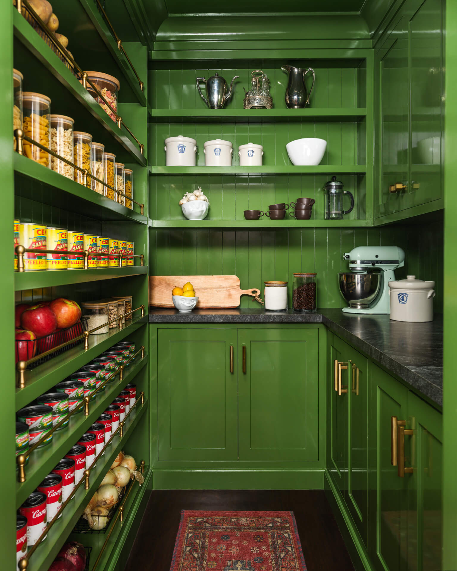 Deep green pantry with open shelves, brass hardware, small appliances, and neatly arranged food storage.