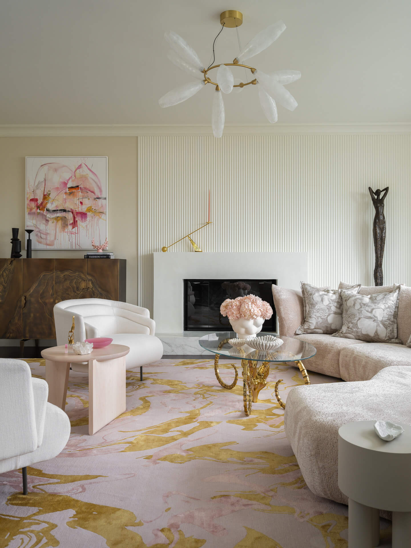 Soft neutral living room with curved seating, fireplace, pastel artwork, floral rug, and sculptural ceiling light.
