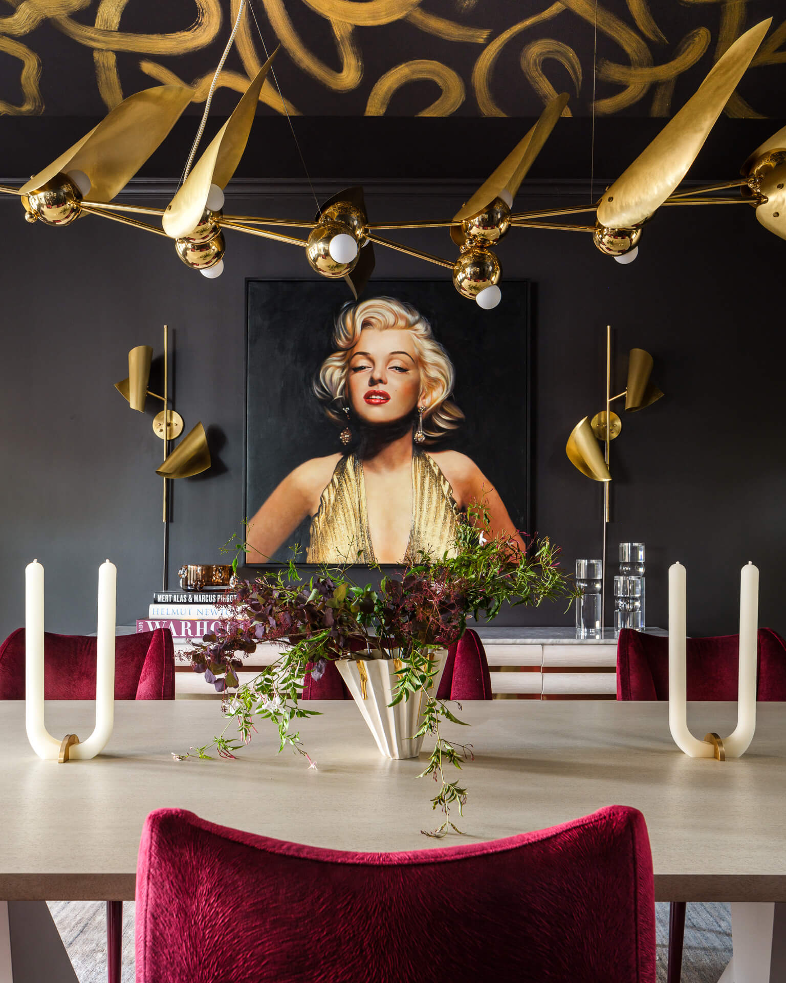 Dramatic dining room with red velvet chairs, black walls, gold chandelier, and a large Marilyn Monroe portrait.