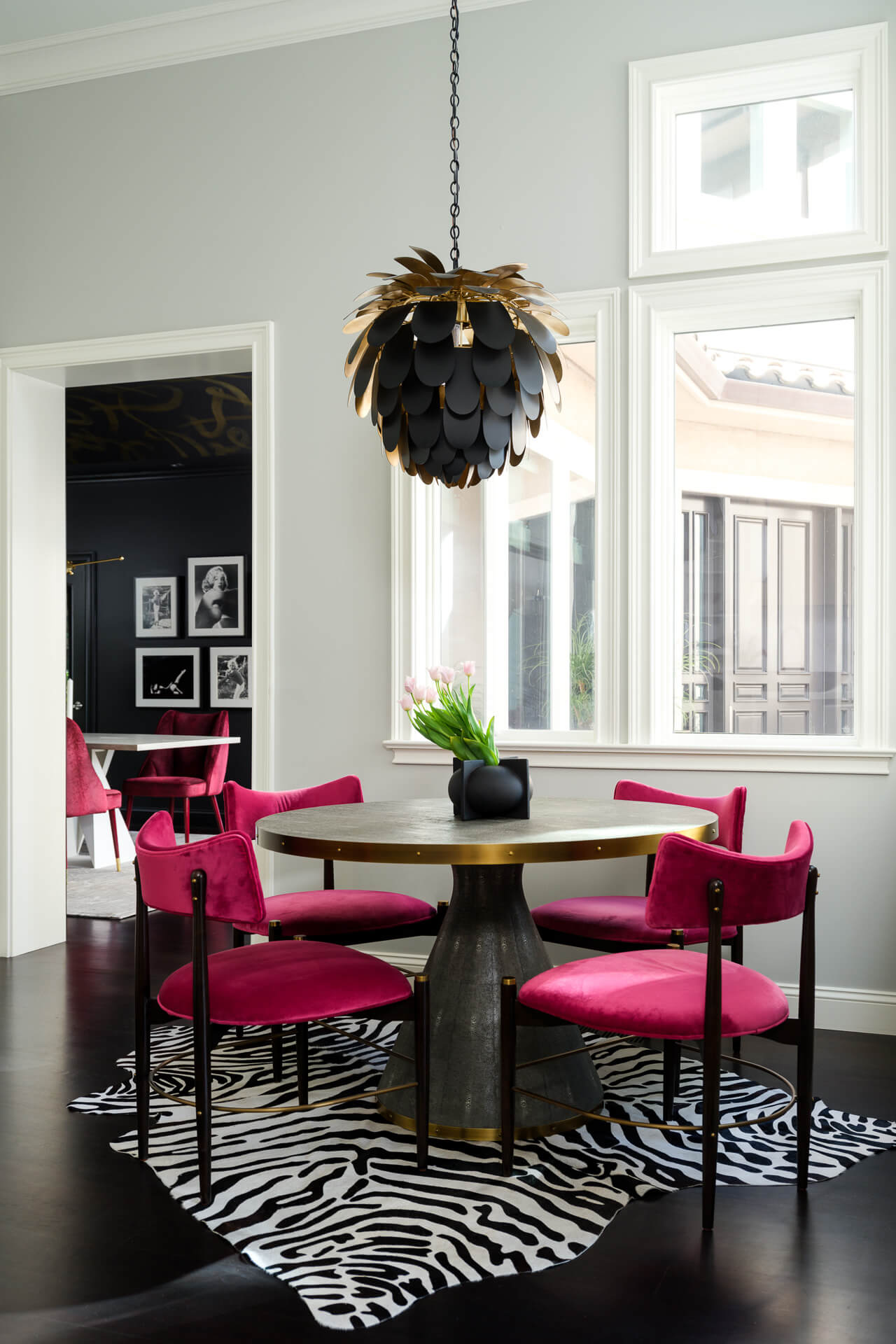 Dining nook with round table, bright pink chairs, zebra-patterned rug, and sculptural black pendant light.