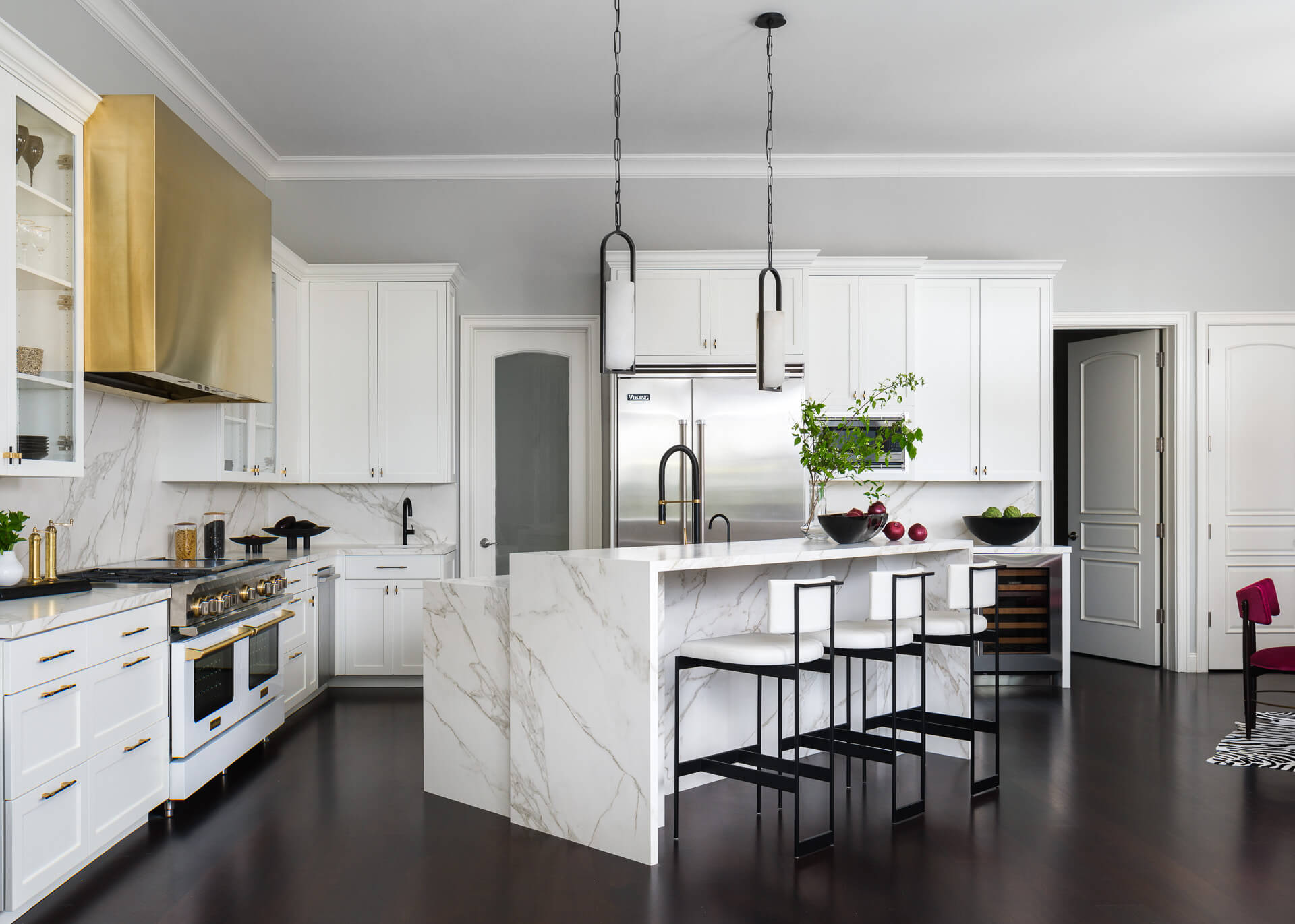 Bright white kitchen with marble island, black stools, brass range hood, and dark wood floors.