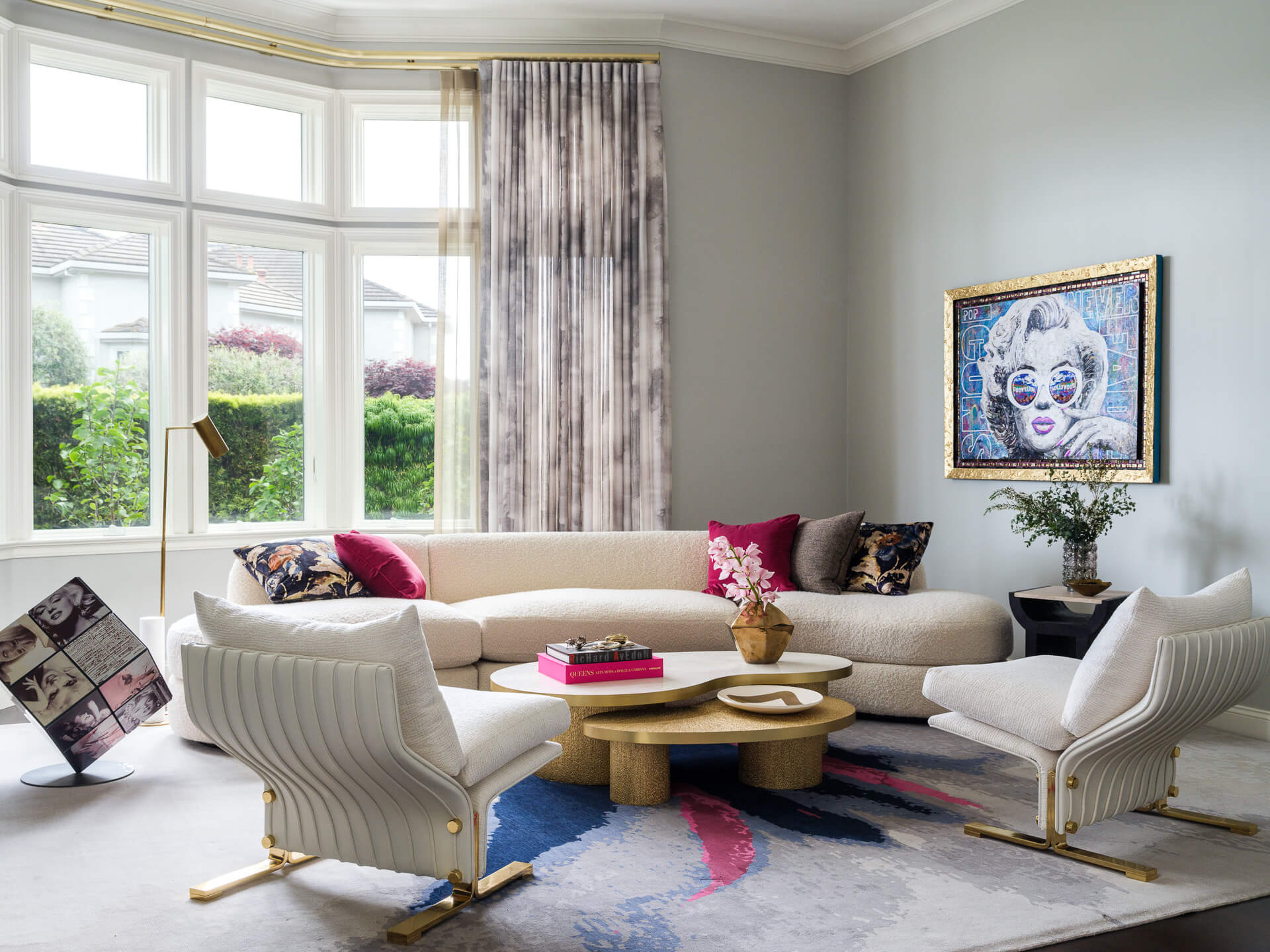 Bright living room with cream sectional, white armchairs, colorful artwork, patterned rug, and large bay windows.