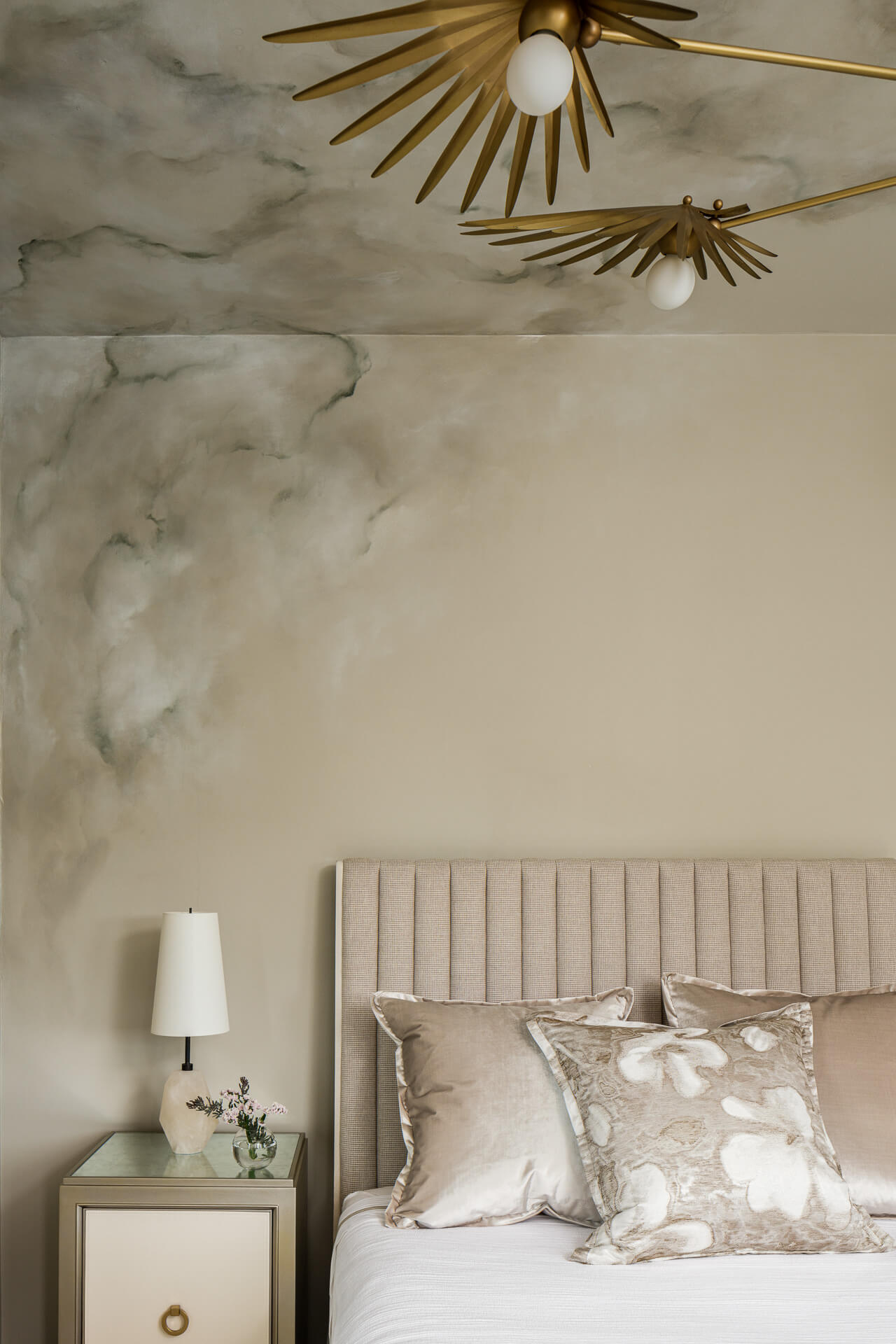 Bedroom detail with upholstered headboard, pale bedding, cloud-like mural wall, and brass starburst ceiling lights.