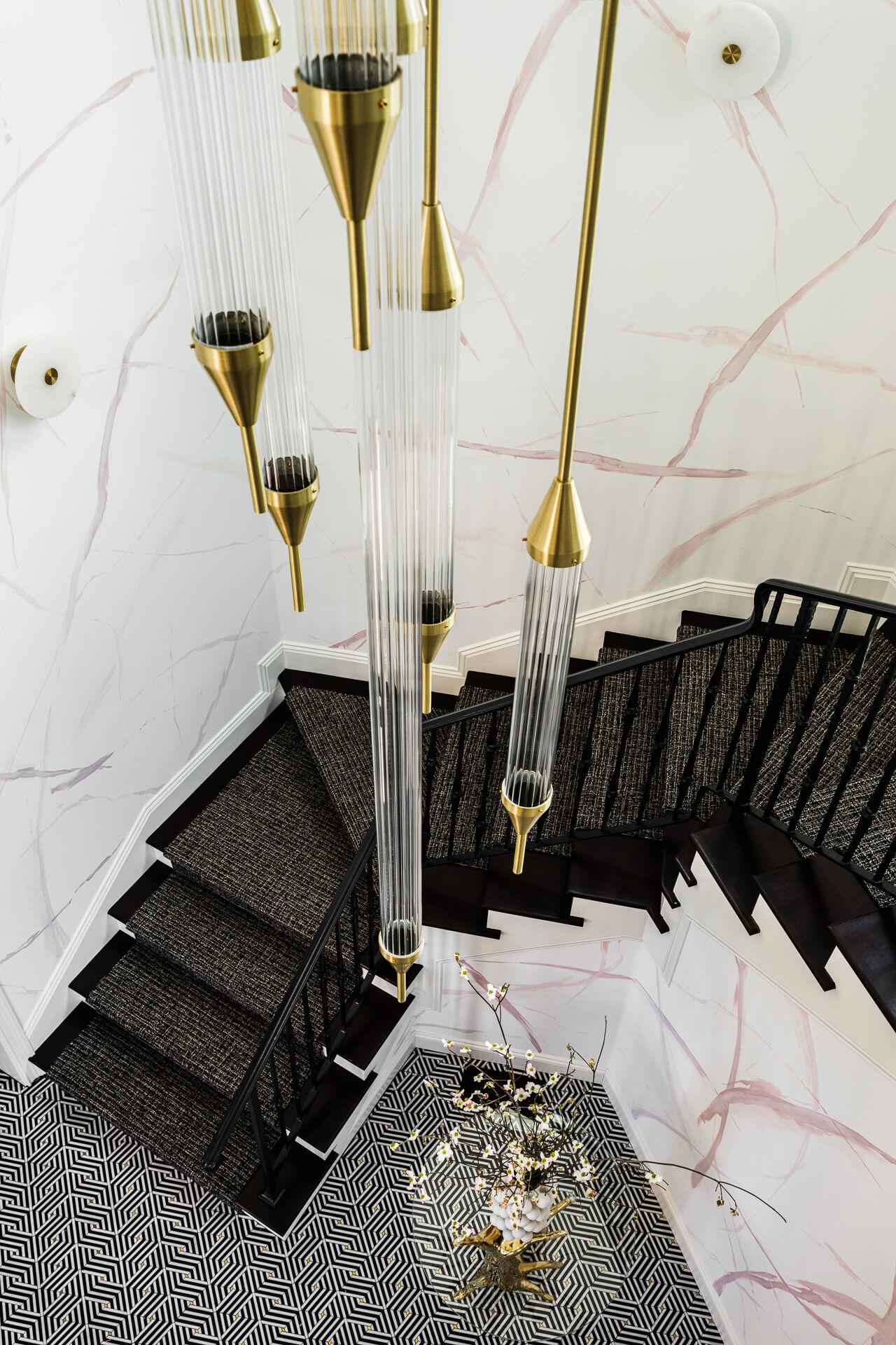 Top-down view of a black-carpeted staircase surrounded by pale marble walls and cascading brass pendant lights.