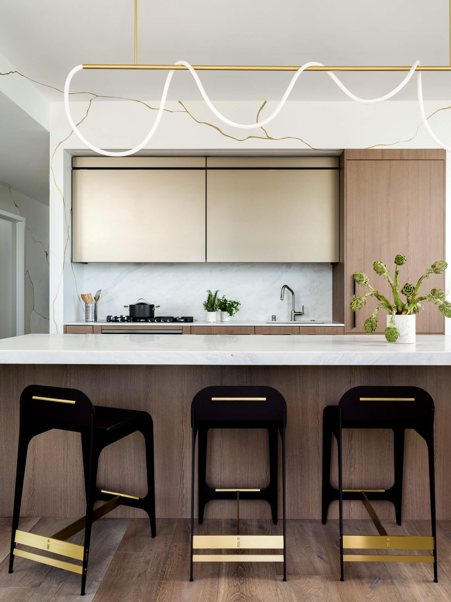 Contemporary kitchen with pale stone island, three black stools, warm wood cabinetry, and sculptural linear lighting.