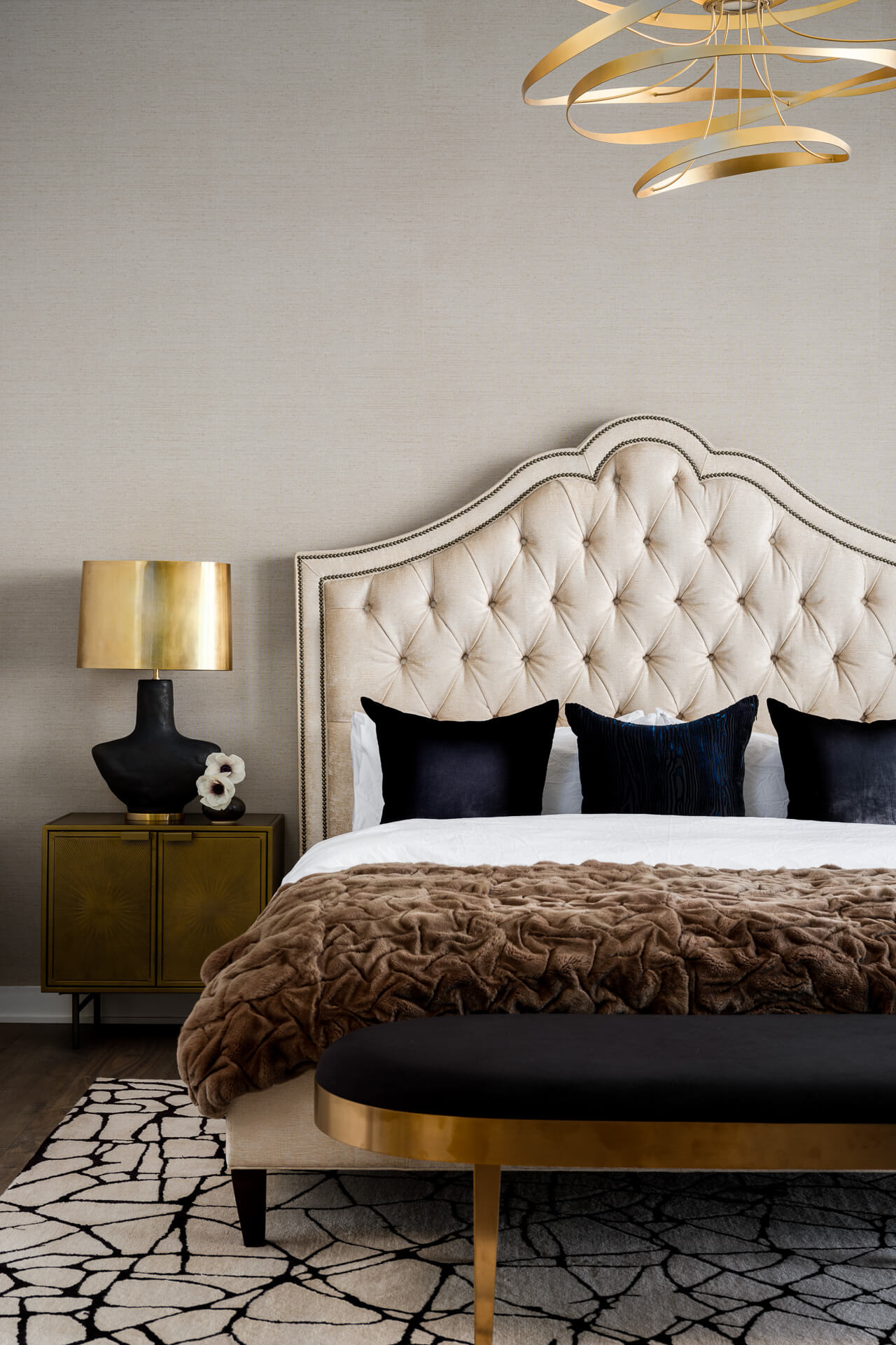 Bedroom with tufted cream headboard, dark accent pillows, textured brown throw, and black-and-gold bench.