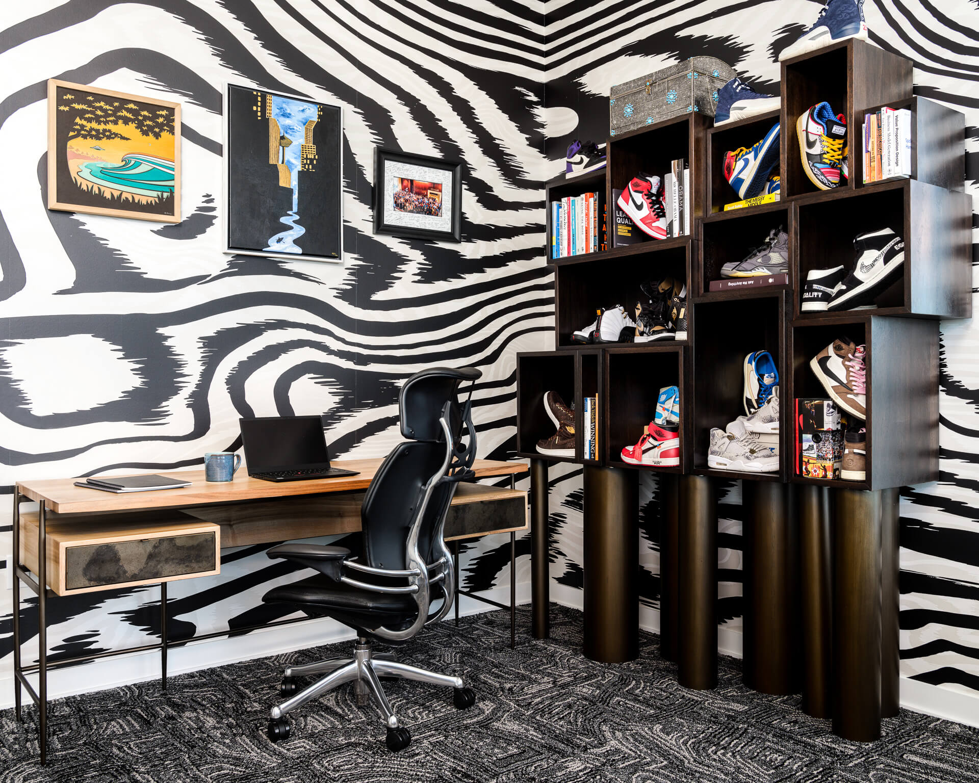 Home office with black-and-white patterned walls, wood desk, black chair, and display shelving filled with art and books.