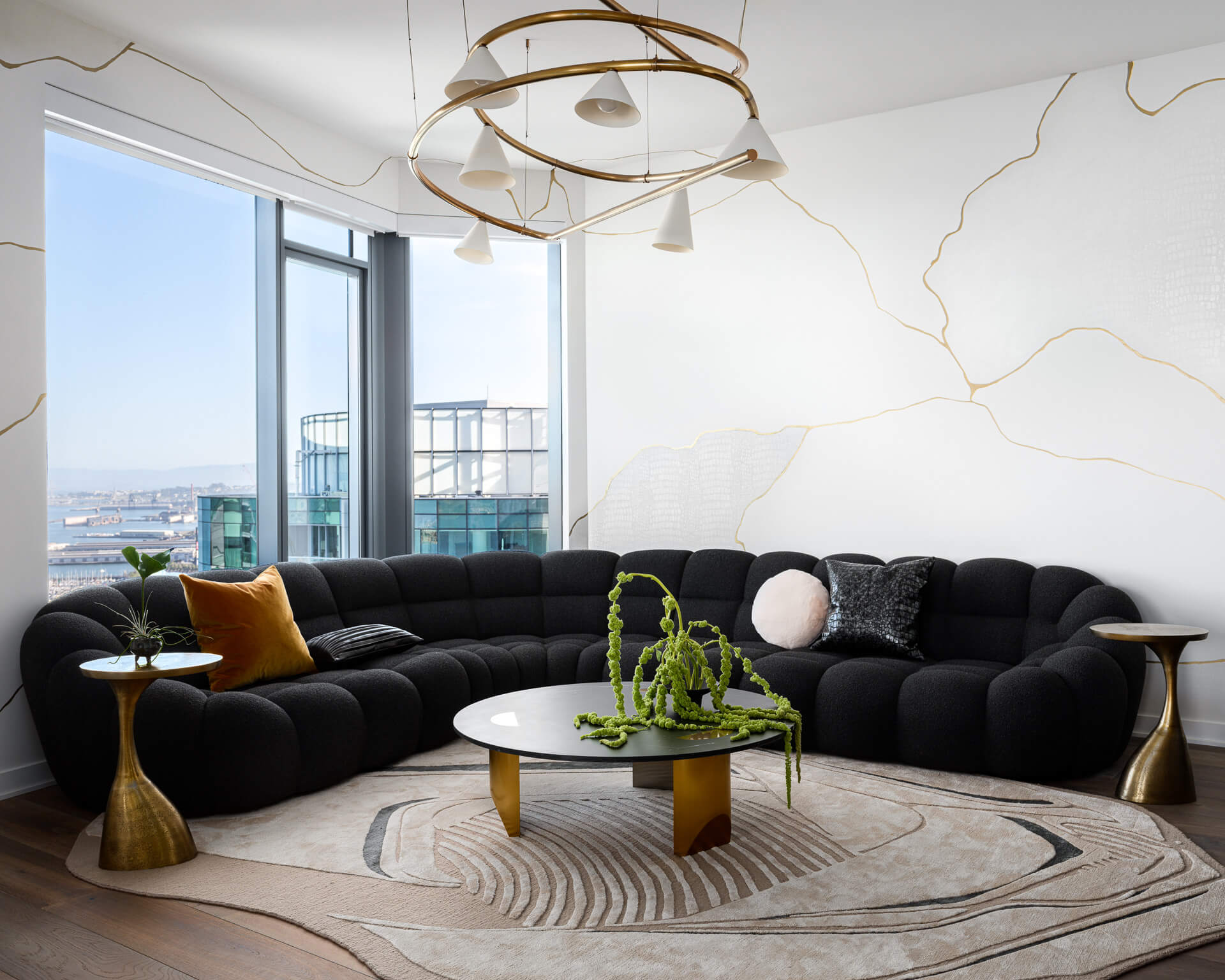 Living room with curved black sectional, round coffee table, sculptural gold chandelier, and panoramic city views.