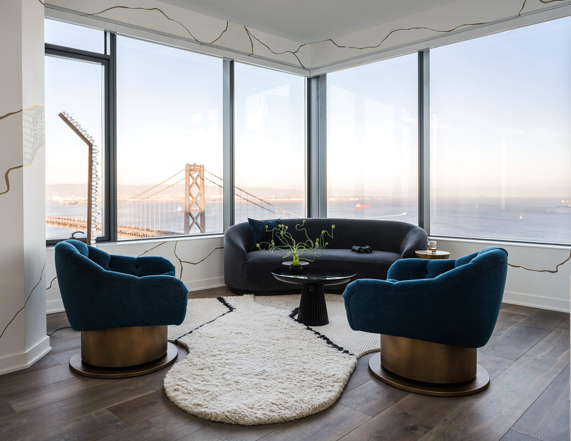 Sitting area with curved black sofa, blue velvet chairs, and expansive views of the Bay Bridge and water.