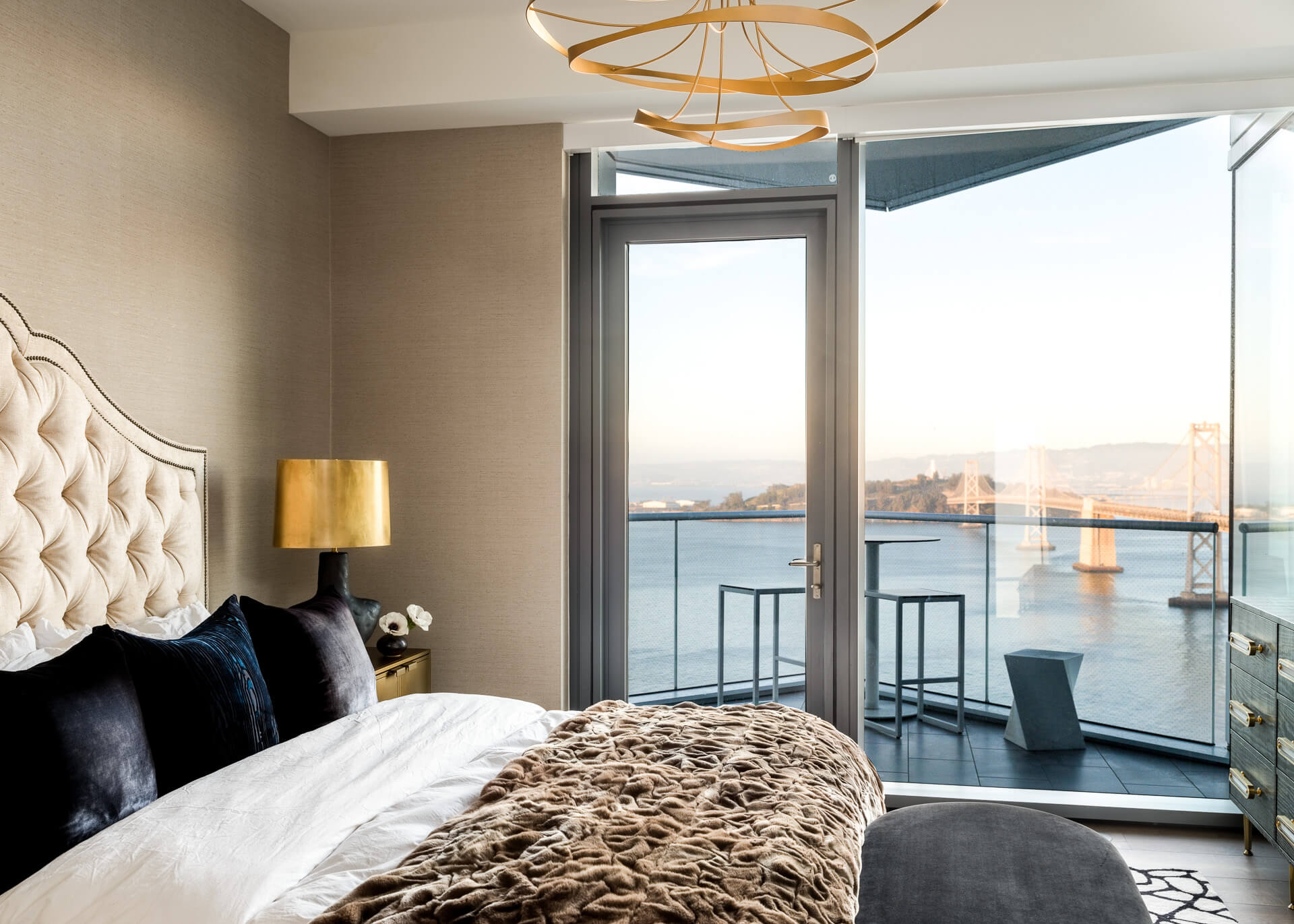 Bedroom with tufted cream headboard, dark pillows, textured throw, and balcony doors overlooking the bay.