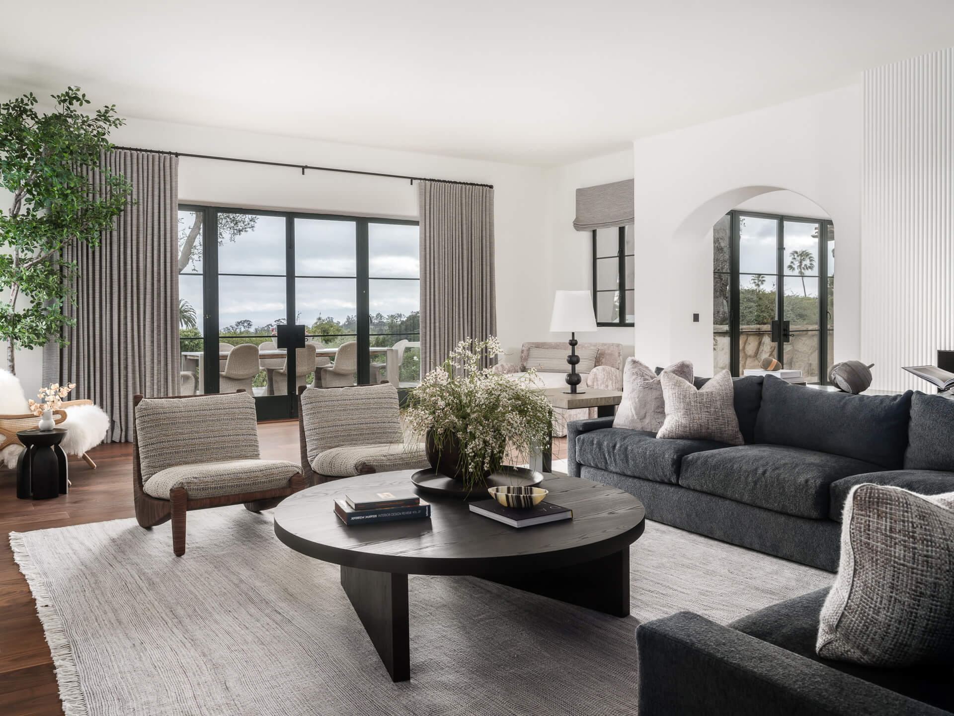 Relaxed living room with charcoal sofa, woven lounge chairs, round black coffee table, and large garden-facing windows.