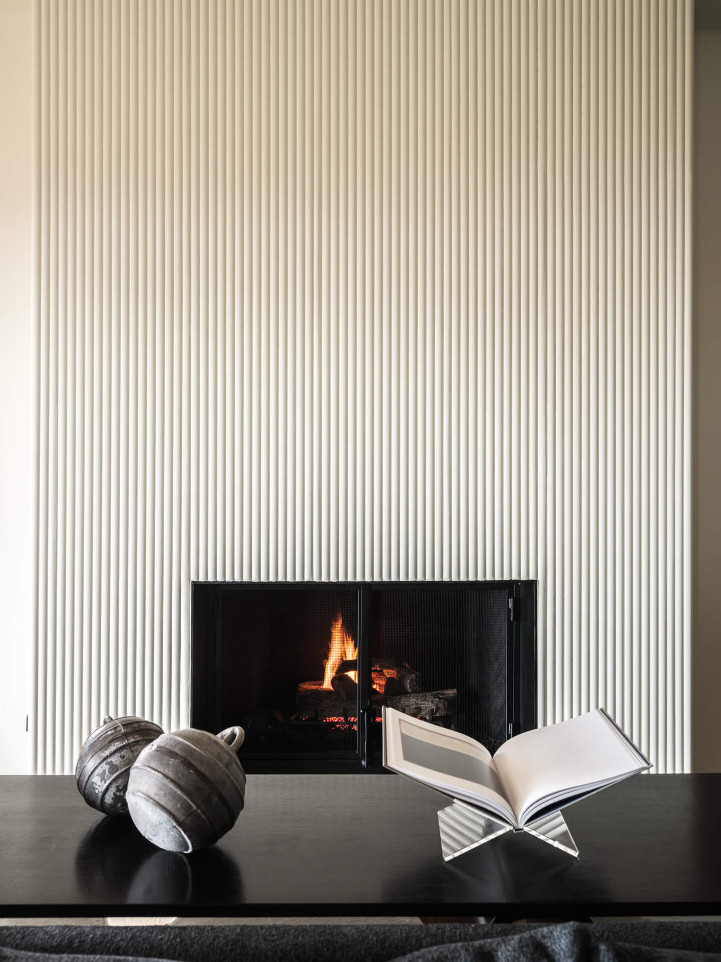 Minimal fireplace set into a vertically ribbed white wall with sculptural objects and an open book in the foreground.
