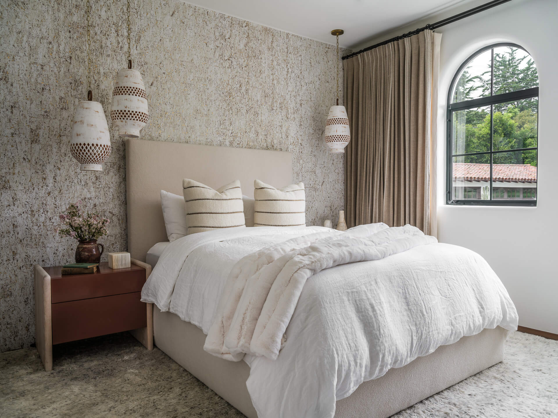 Neutral bedroom with upholstered bed, textured wall covering, woven pendant lights, and tall arched window.
