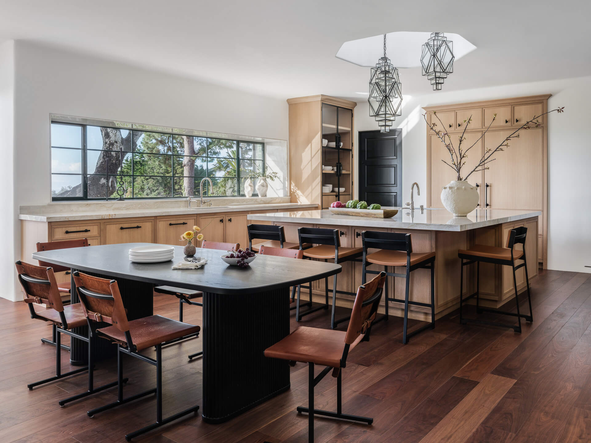 Open kitchen and dining area with wood cabinetry, dark island, leather chairs, and large garden-facing windows.