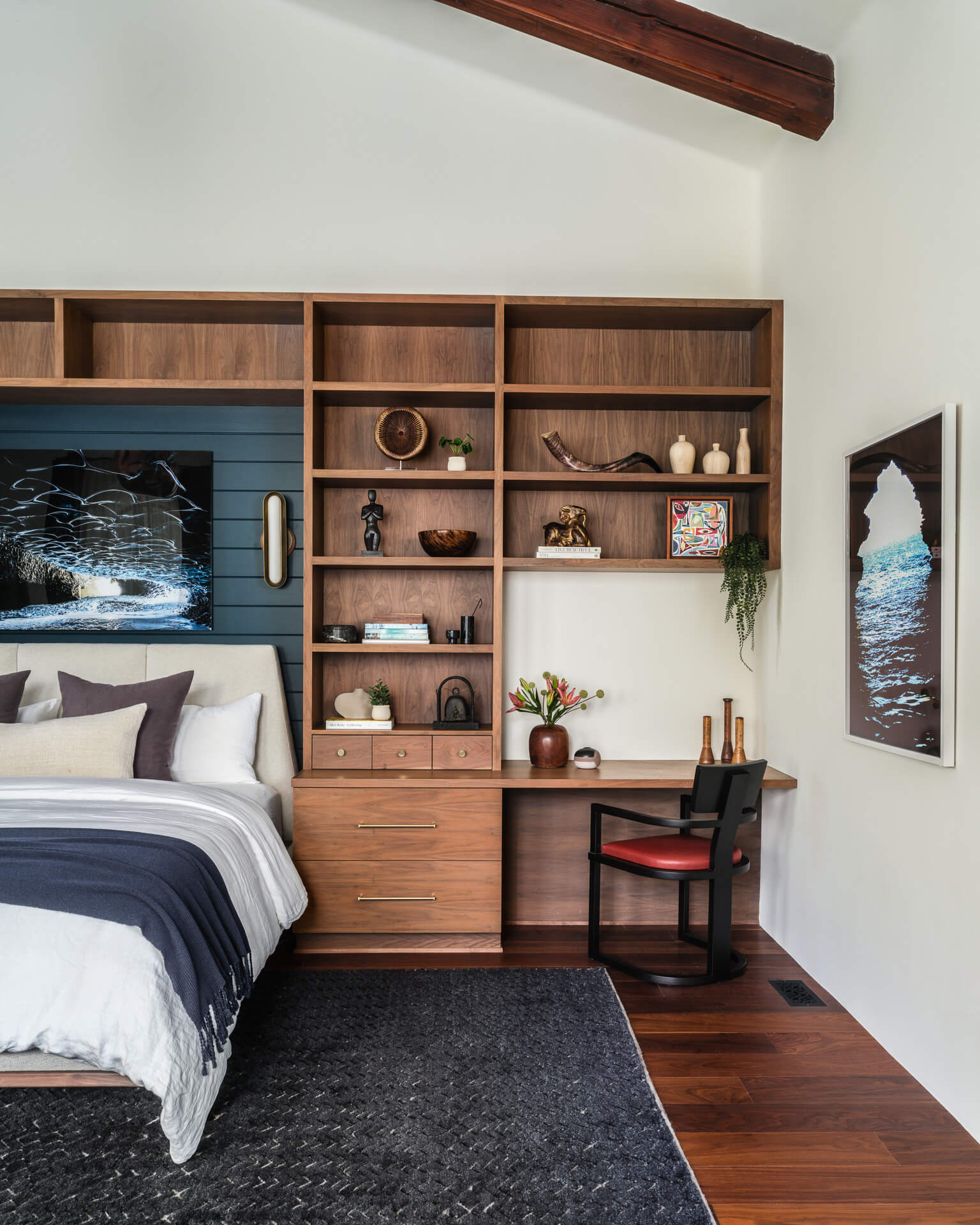 Bedroom with built-in wood shelving and desk, dark bedding, colorful artwork, and exposed ceiling beam.