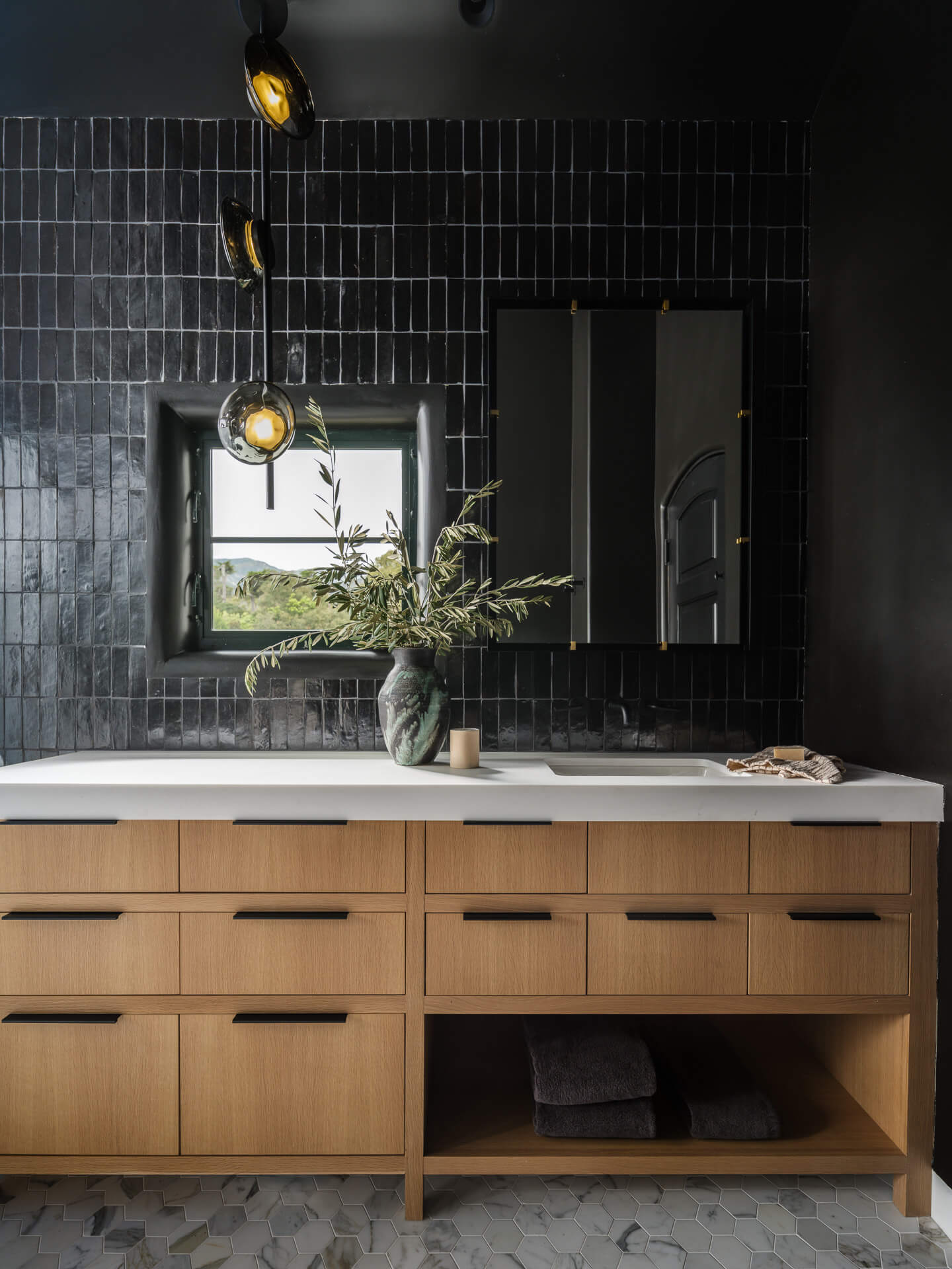 Bathroom vanity with light wood cabinetry, white countertop, dark tiled wall, and large mirror.