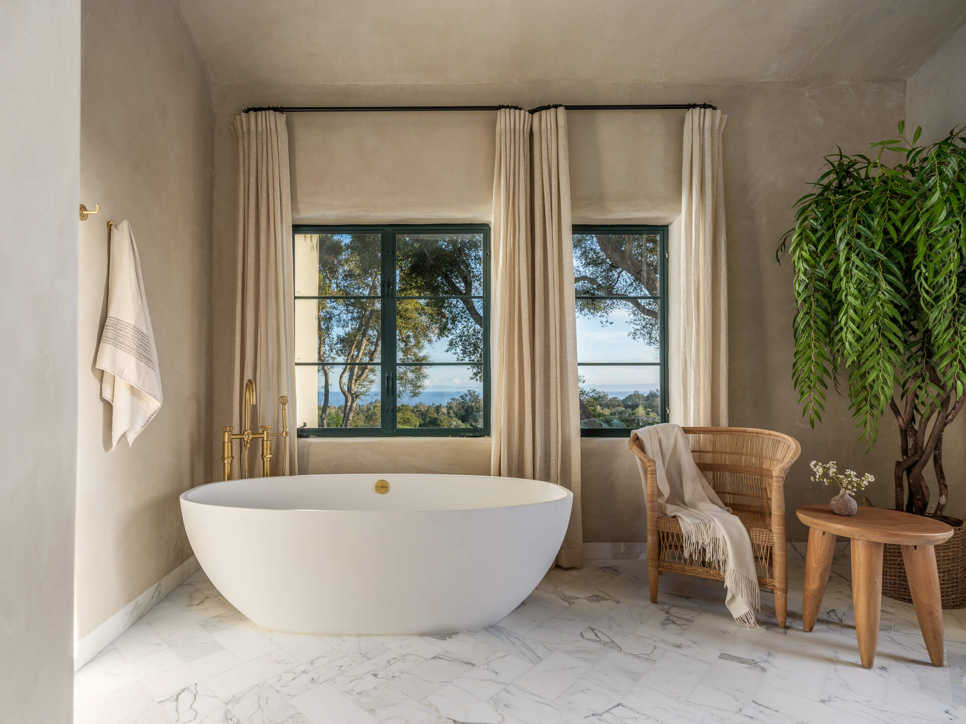 Bright bathroom with freestanding white tub, tall curtained windows, woven chair, and large potted fern.