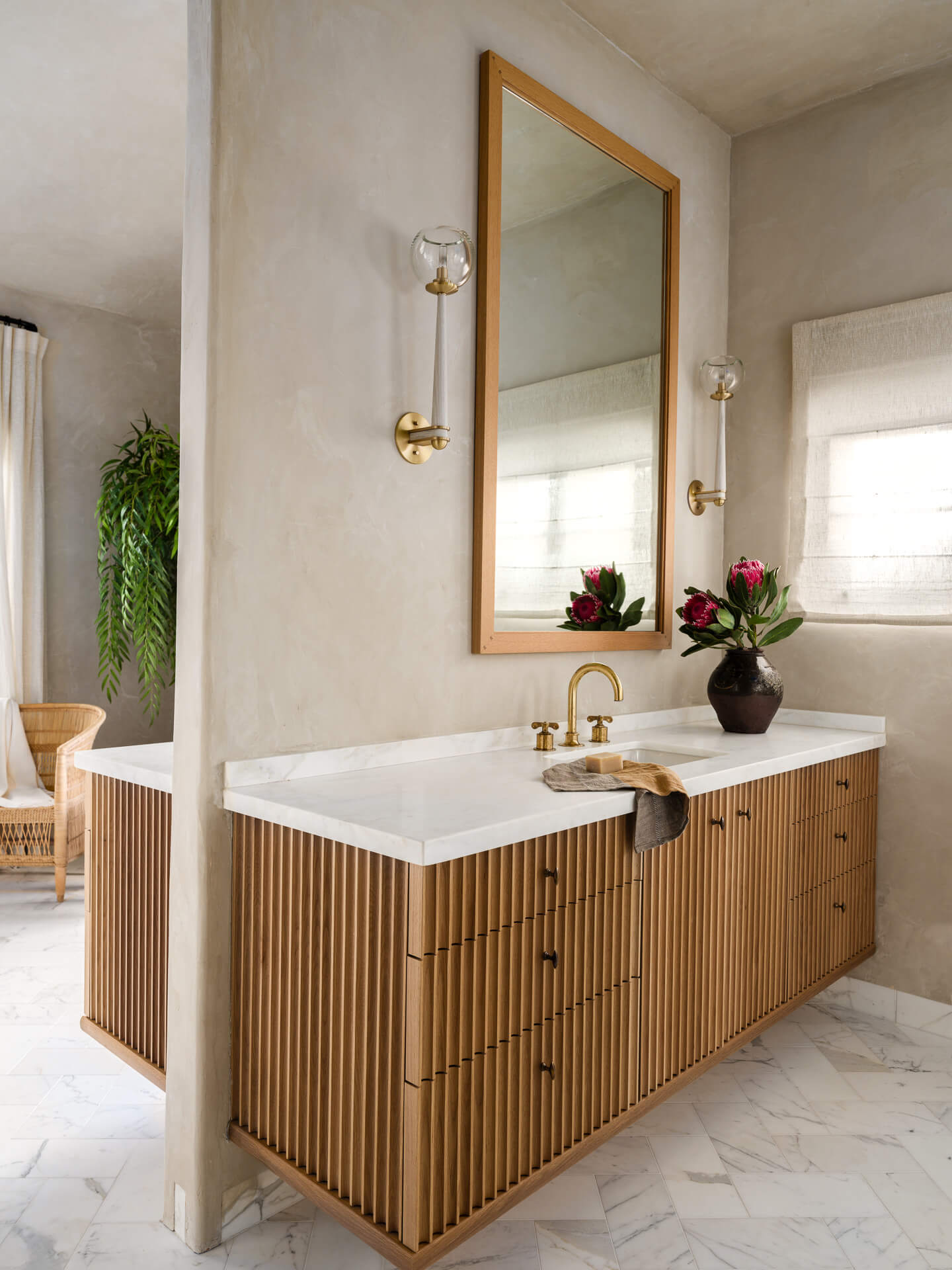 Light wood bathroom vanity with white countertop, brass fixtures, framed mirror, and flowers.