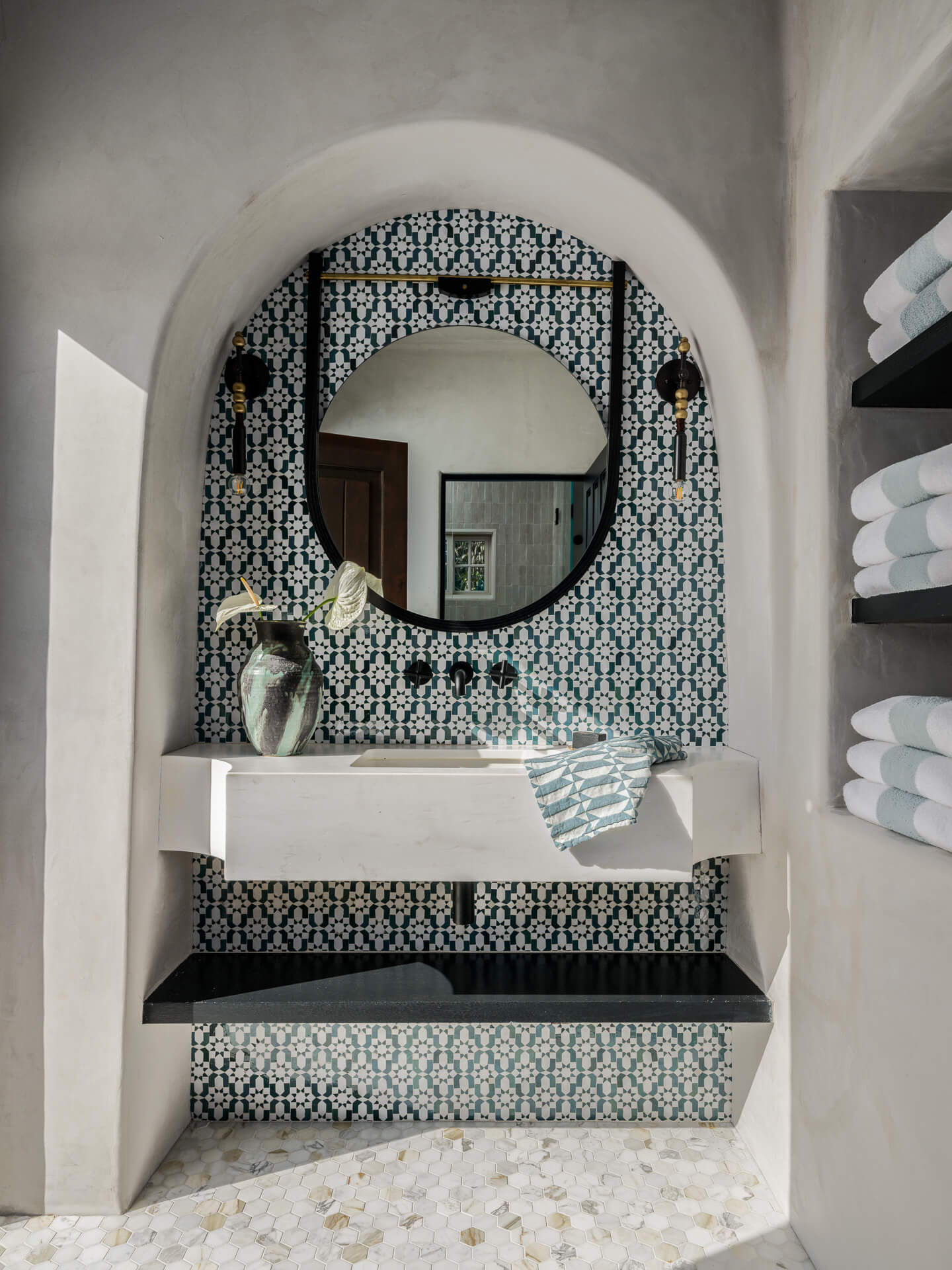 Arched bathroom niche with patterned blue tile, white sink, round mirror, and open towel shelving.