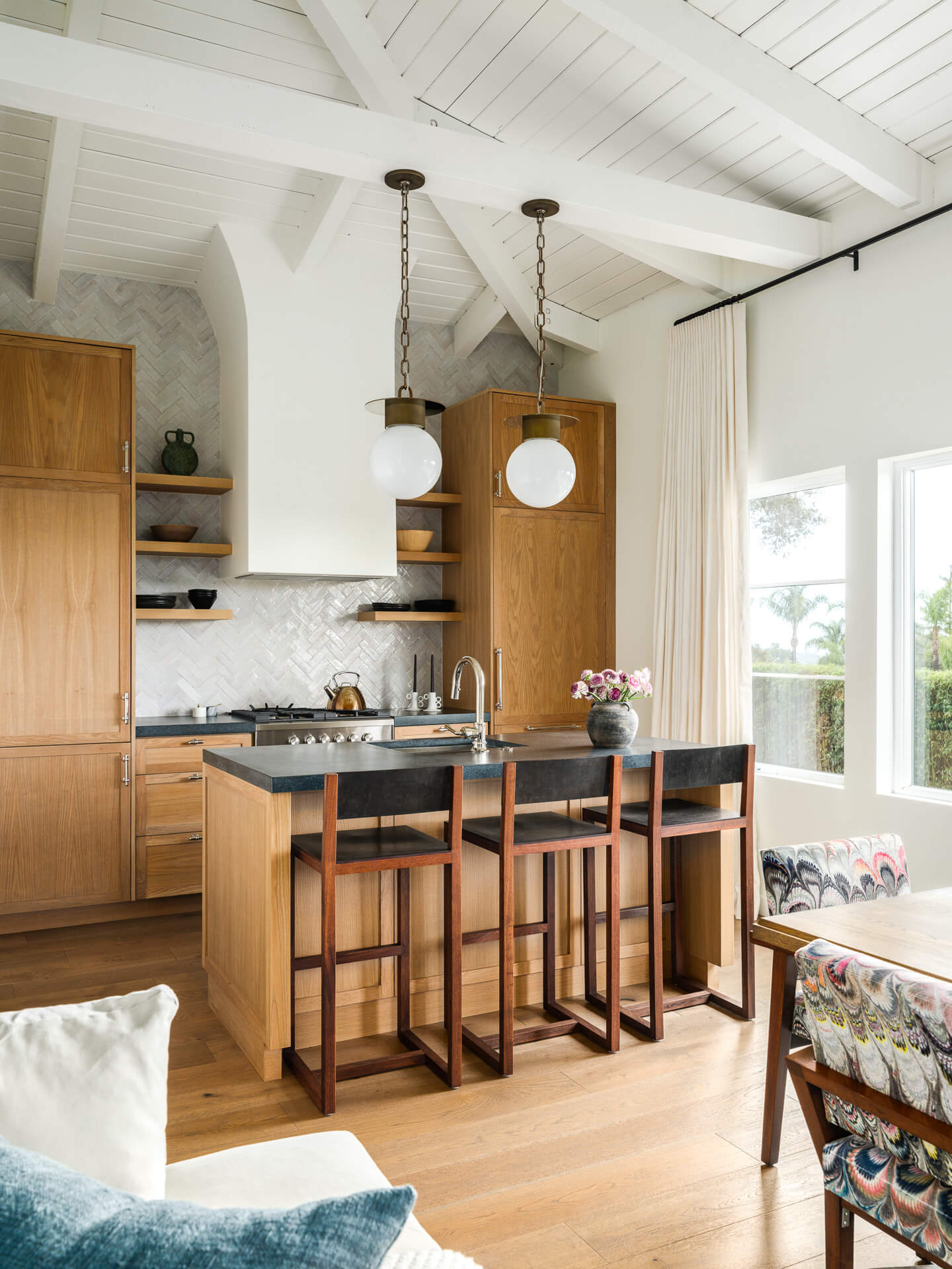 Bright kitchen with wood cabinetry, dark island, globe pendant lights, and white beamed ceiling.