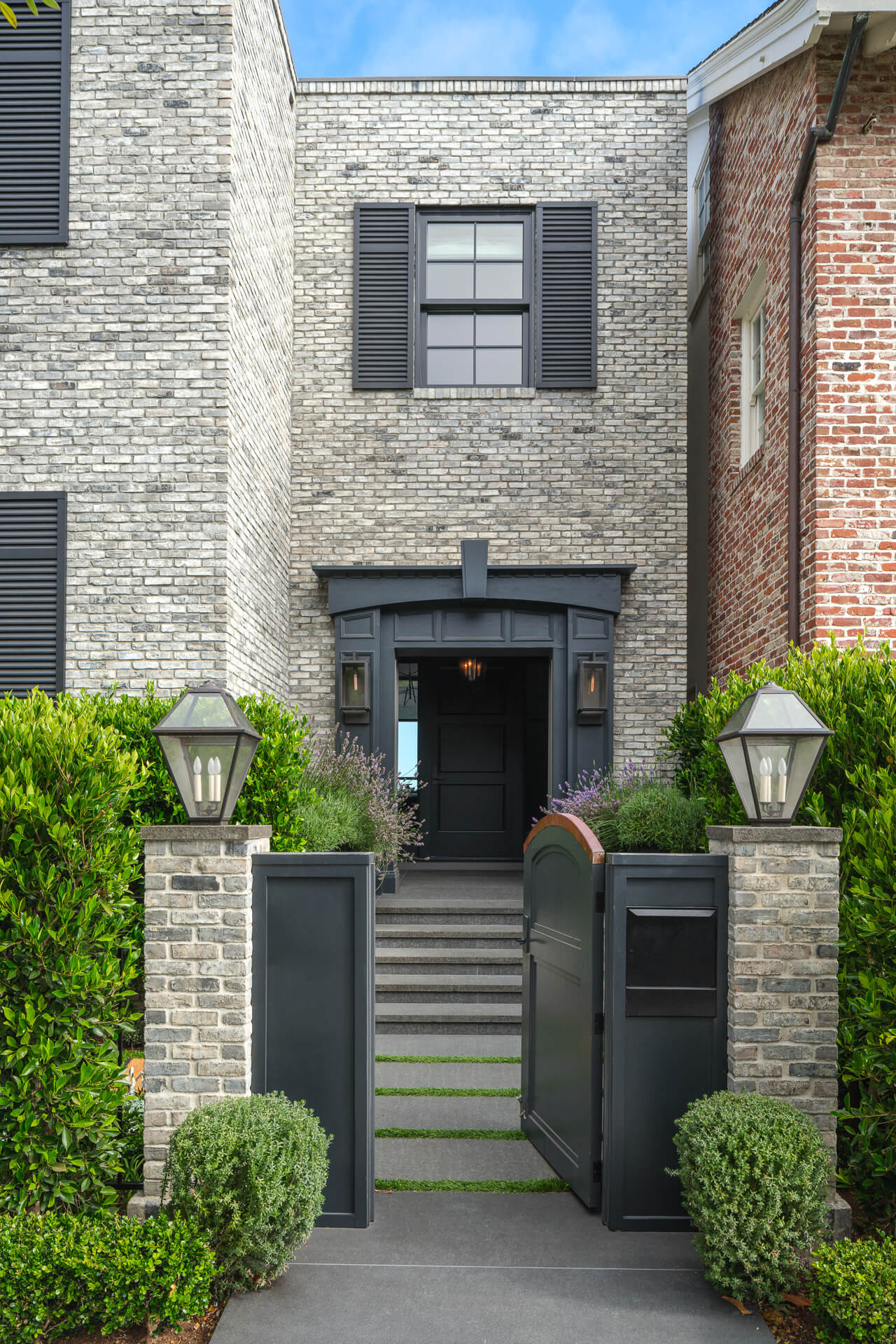 Gray brick townhouse with black shutters, landscaped hedges, and a gated front entrance.