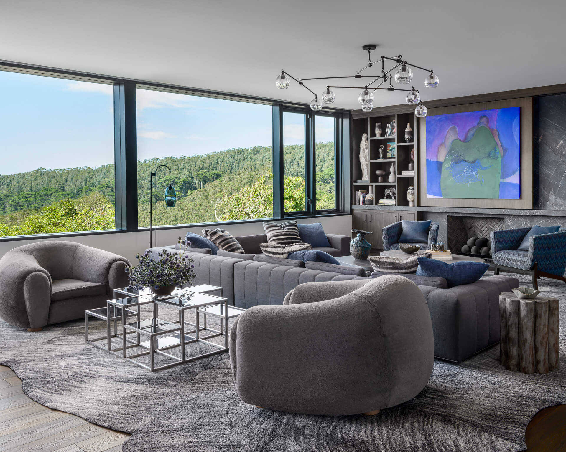 Contemporary living room with curved gray seating, layered tables, large artwork, and panoramic hillside views.