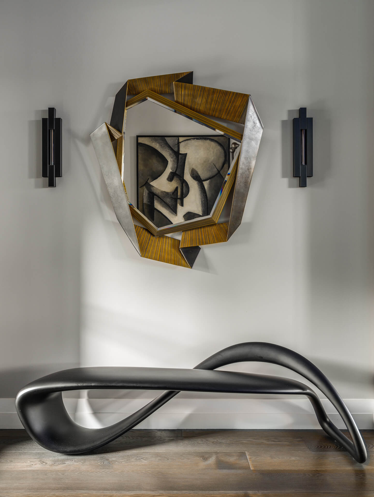Sculptural black bench beneath an angular faceted mirror flanked by dark wall sconces.
