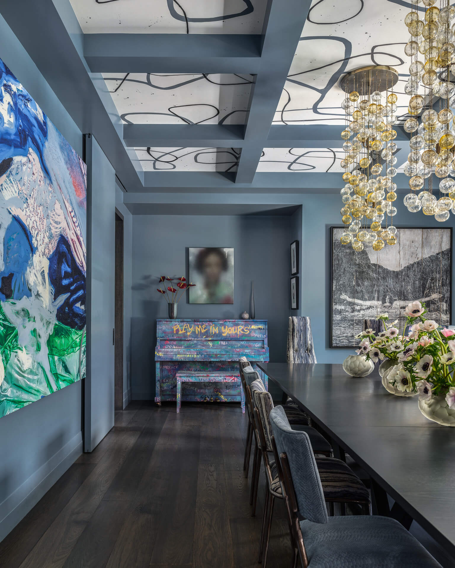 Long dining room with dark table, blue ceiling beams, colorful artwork, and cascading glass pendant lights.