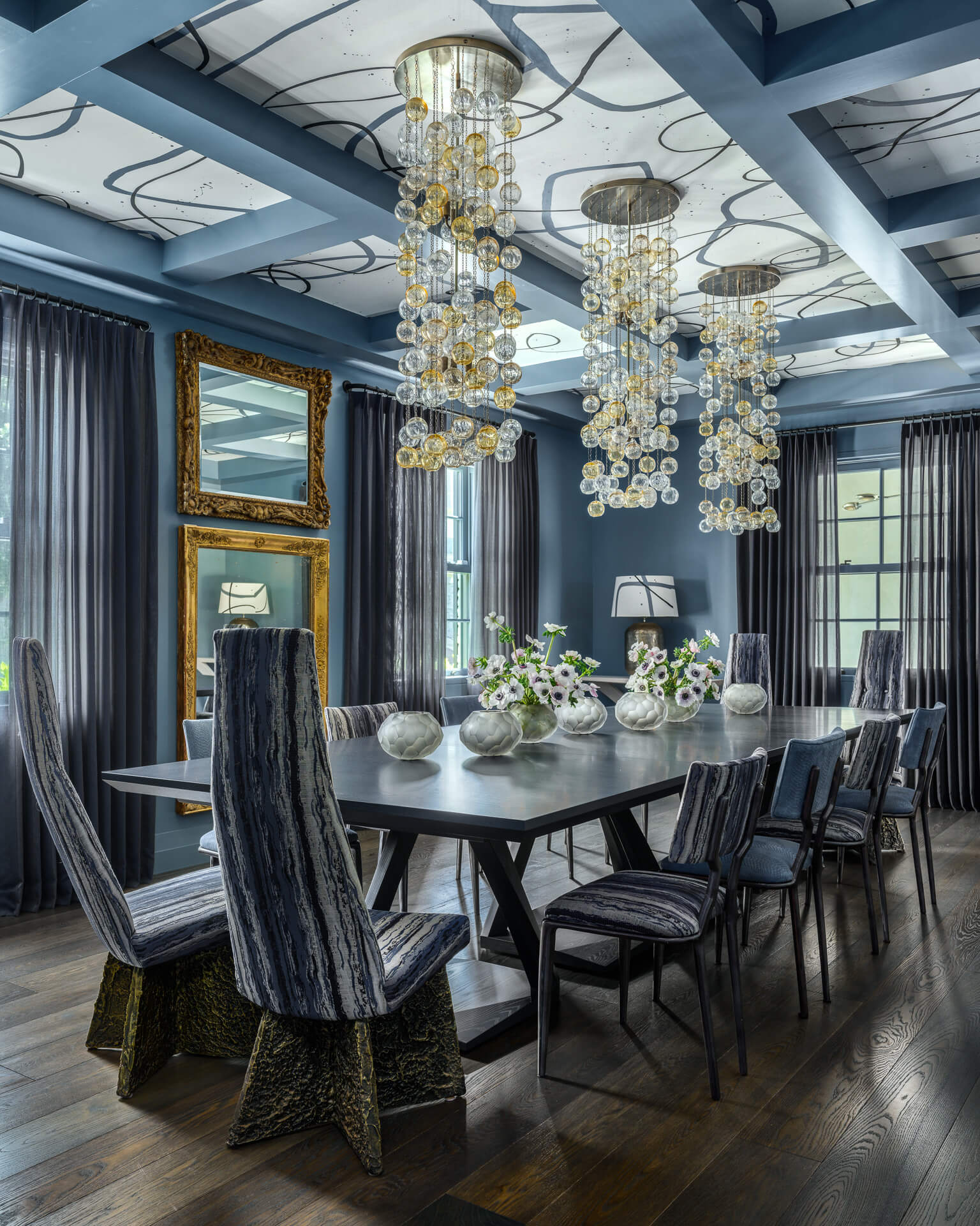 Moody dining room with dark wood table, upholstered chairs, blue-gray walls, and cascading glass chandeliers.